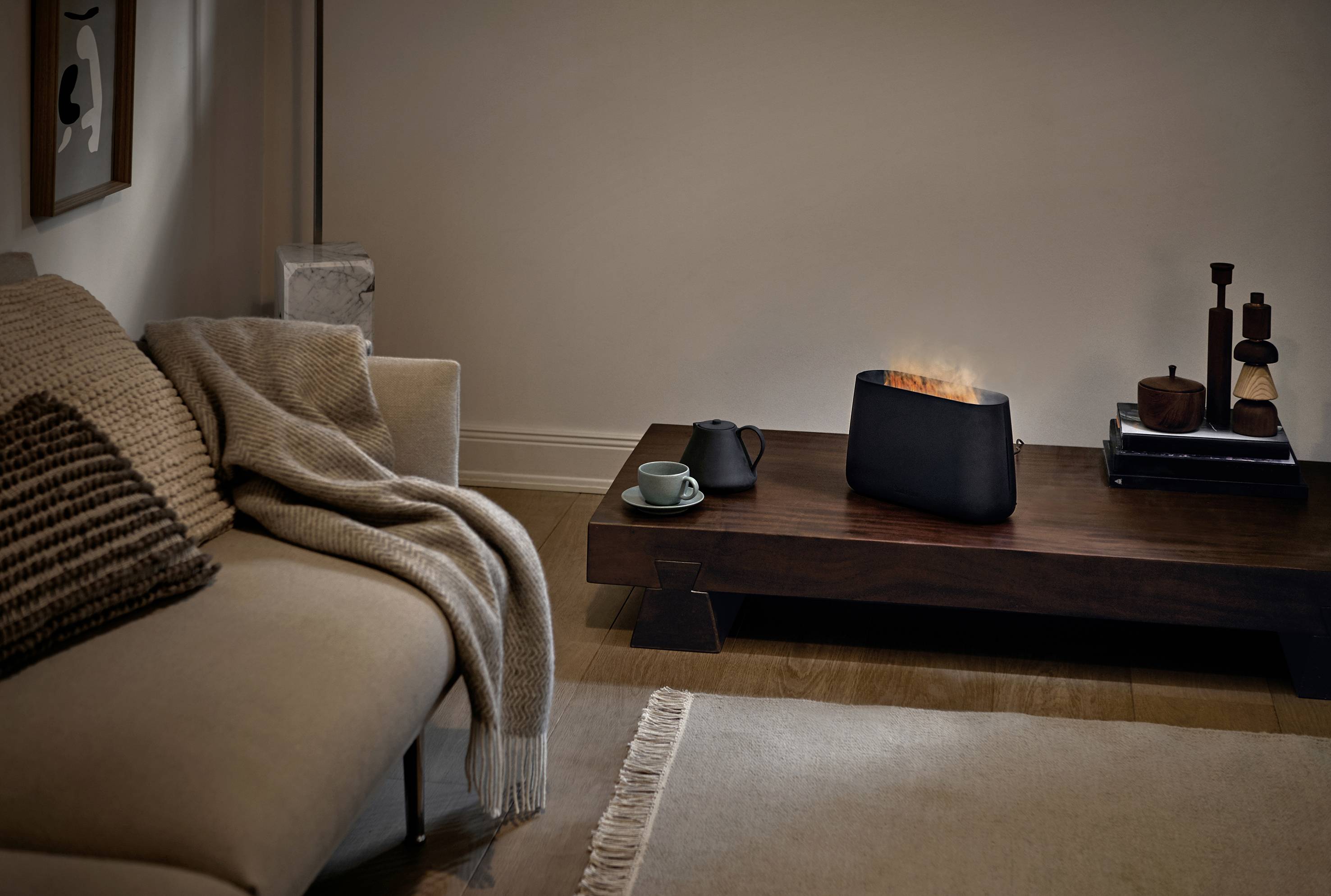 Living room with a beige sofa and throw, wooden coffee table with a tea cup and artificial fireplace, warm lighting.