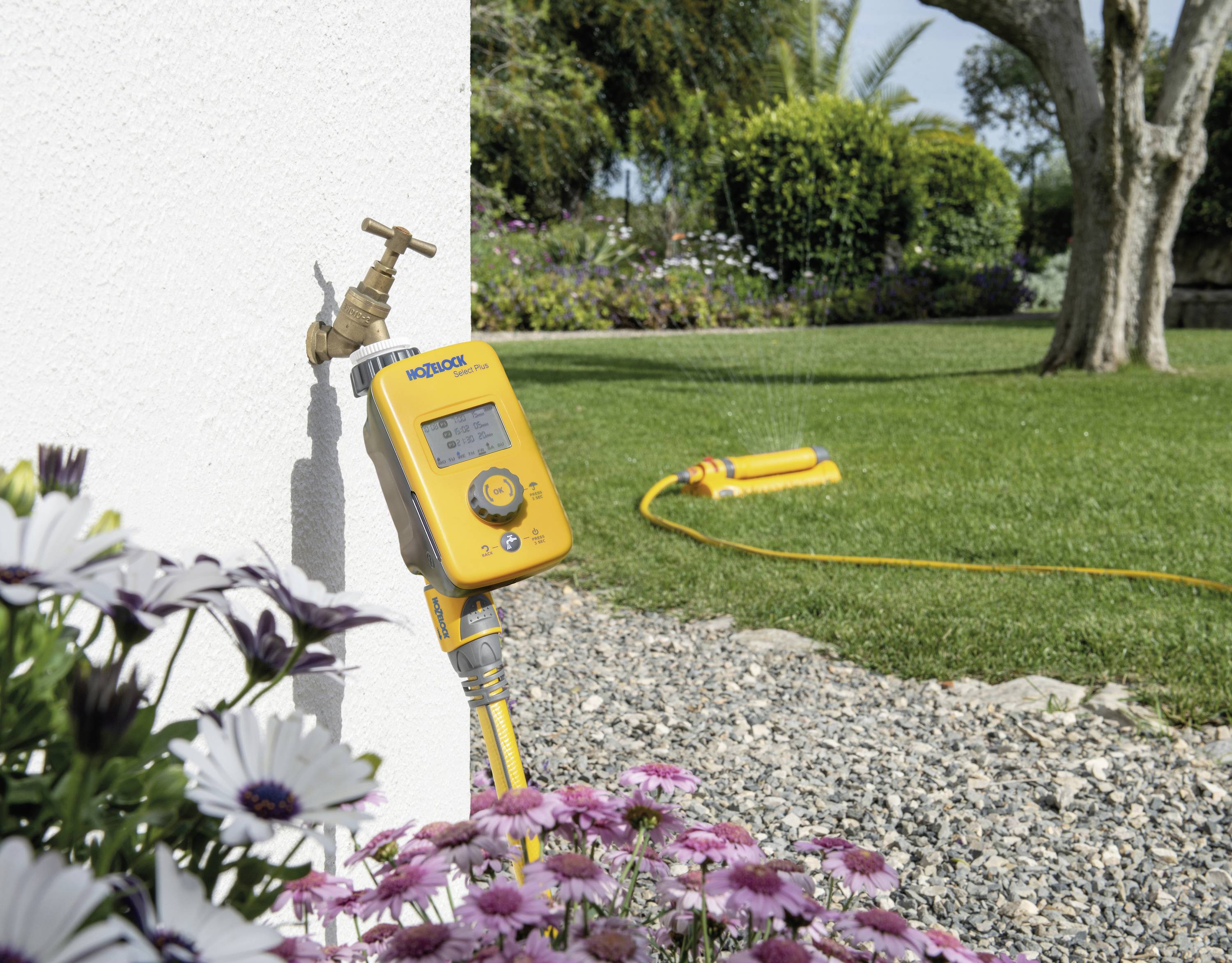 A yellow garden irrigation timer is connected to a tap, with a lawn sprinkler running in a garden in the background.