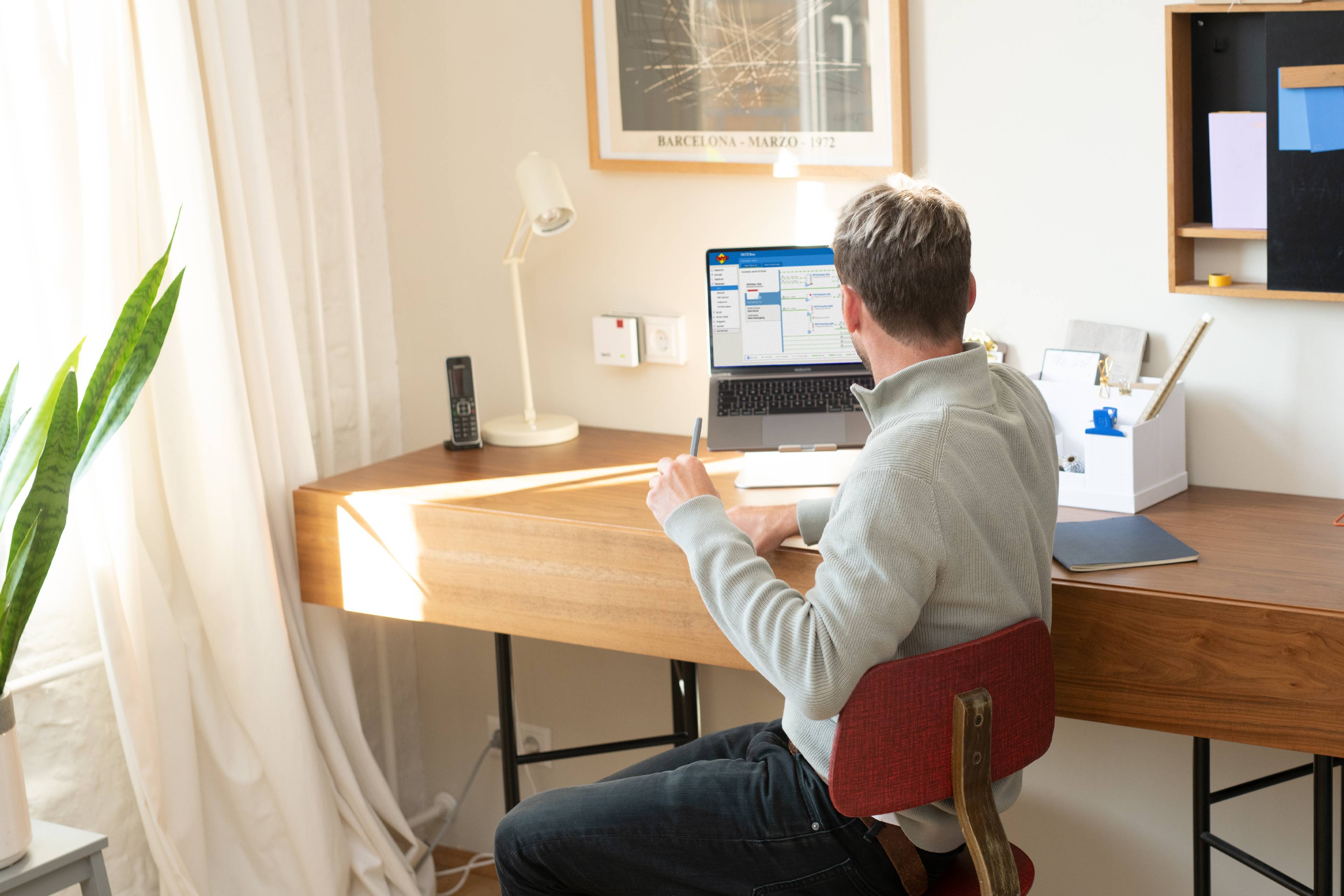 A man is sitting at a desk and working on a laptop. The room is brightly lit, with plants and office supplies around him.