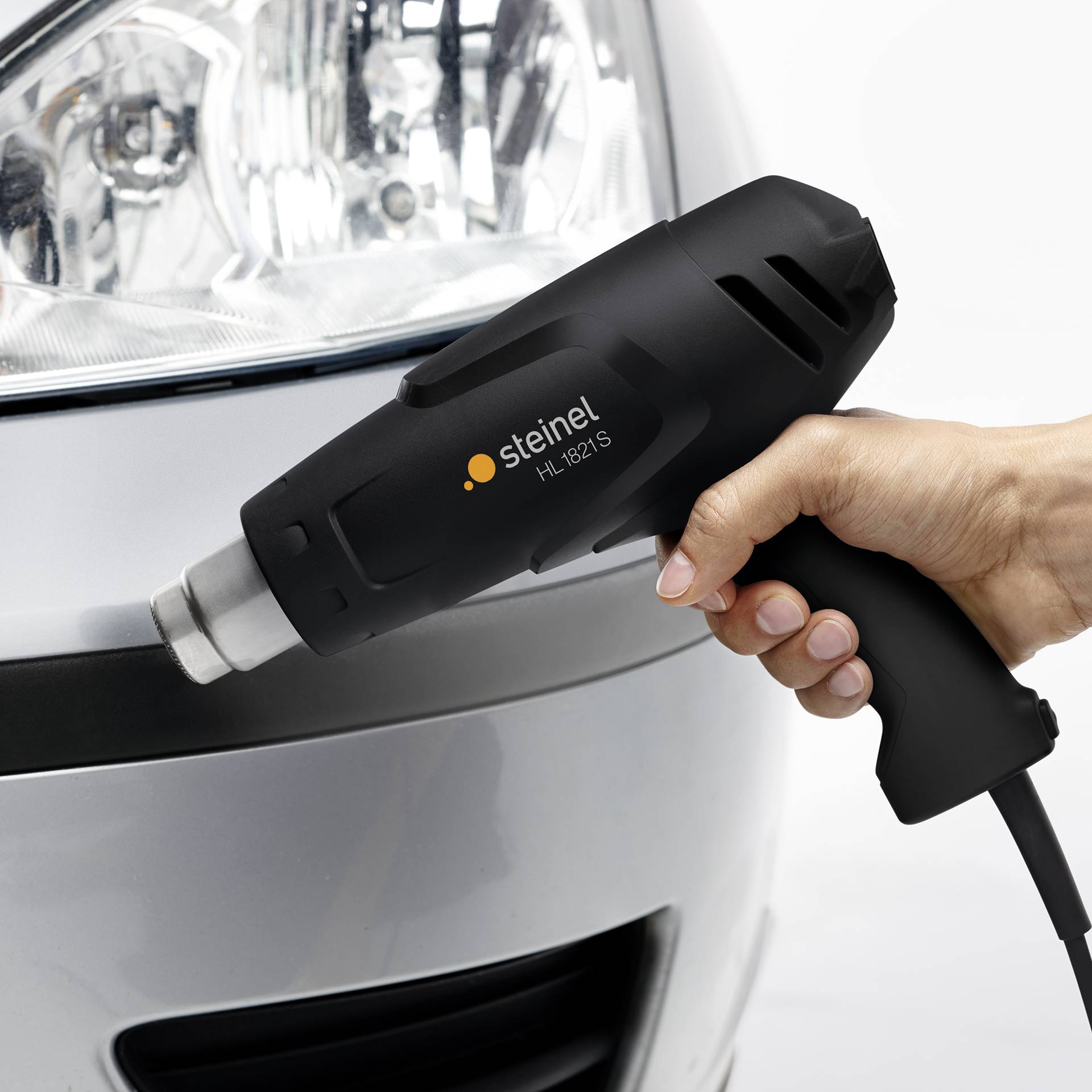A hand is holding a Steinel hot air blower in front of a car headlight. The device is being used for repair or tuning work.