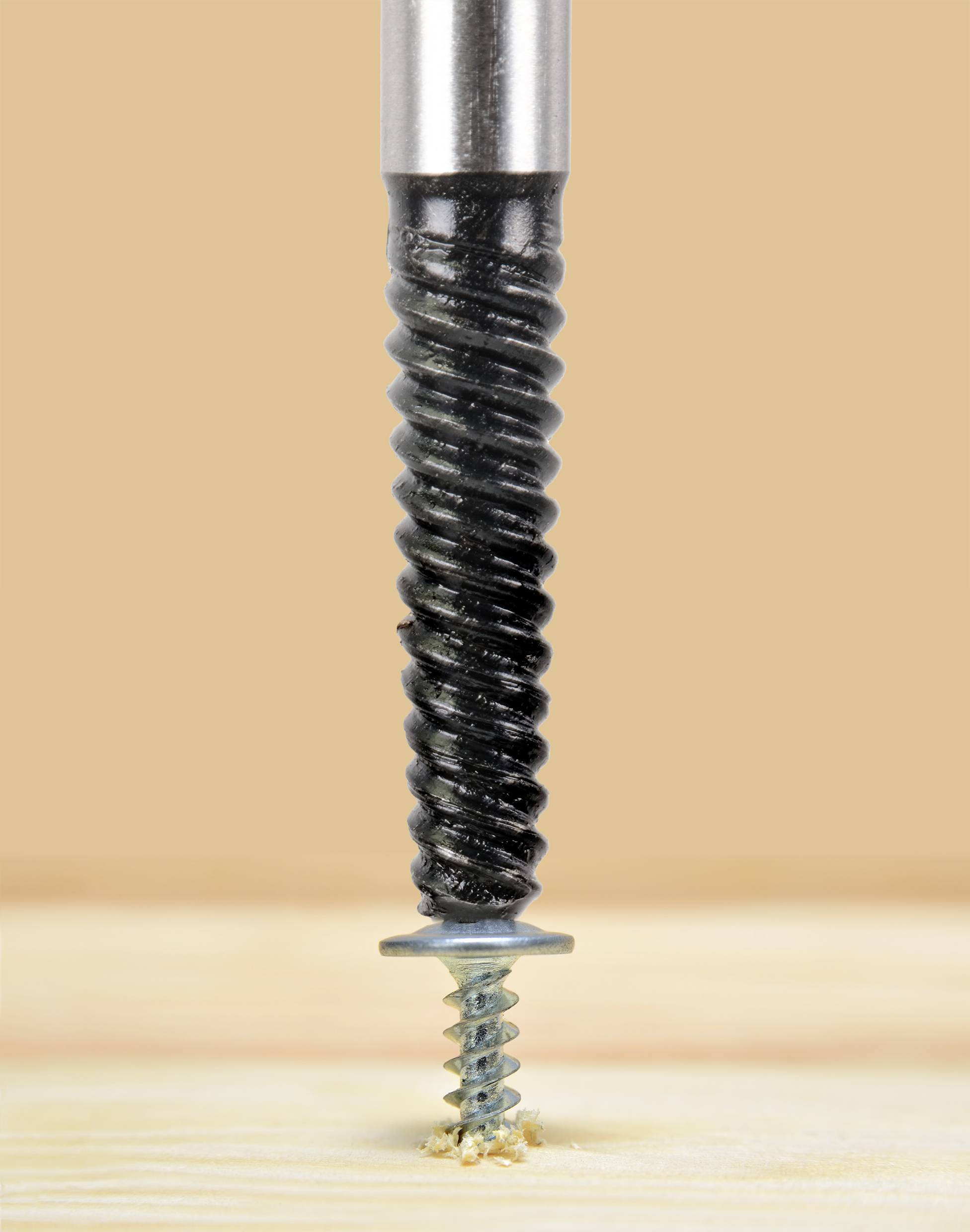 A black drill is driving a silver screw into a wooden board, against a light brown background.