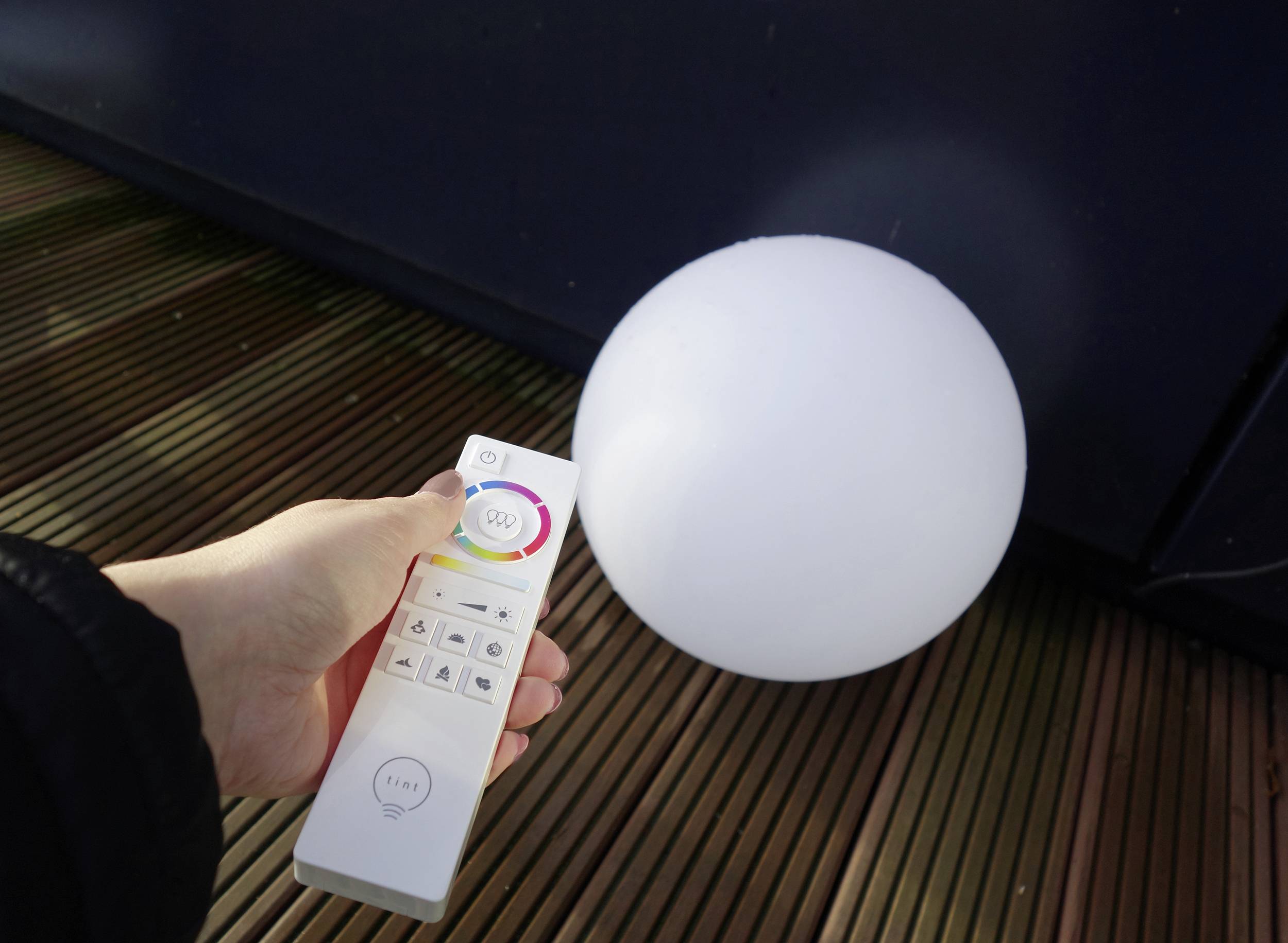 A hand holds a remote control with a colour wheel and buttons, pointing it at a white, round light fixture on a wooden floor.