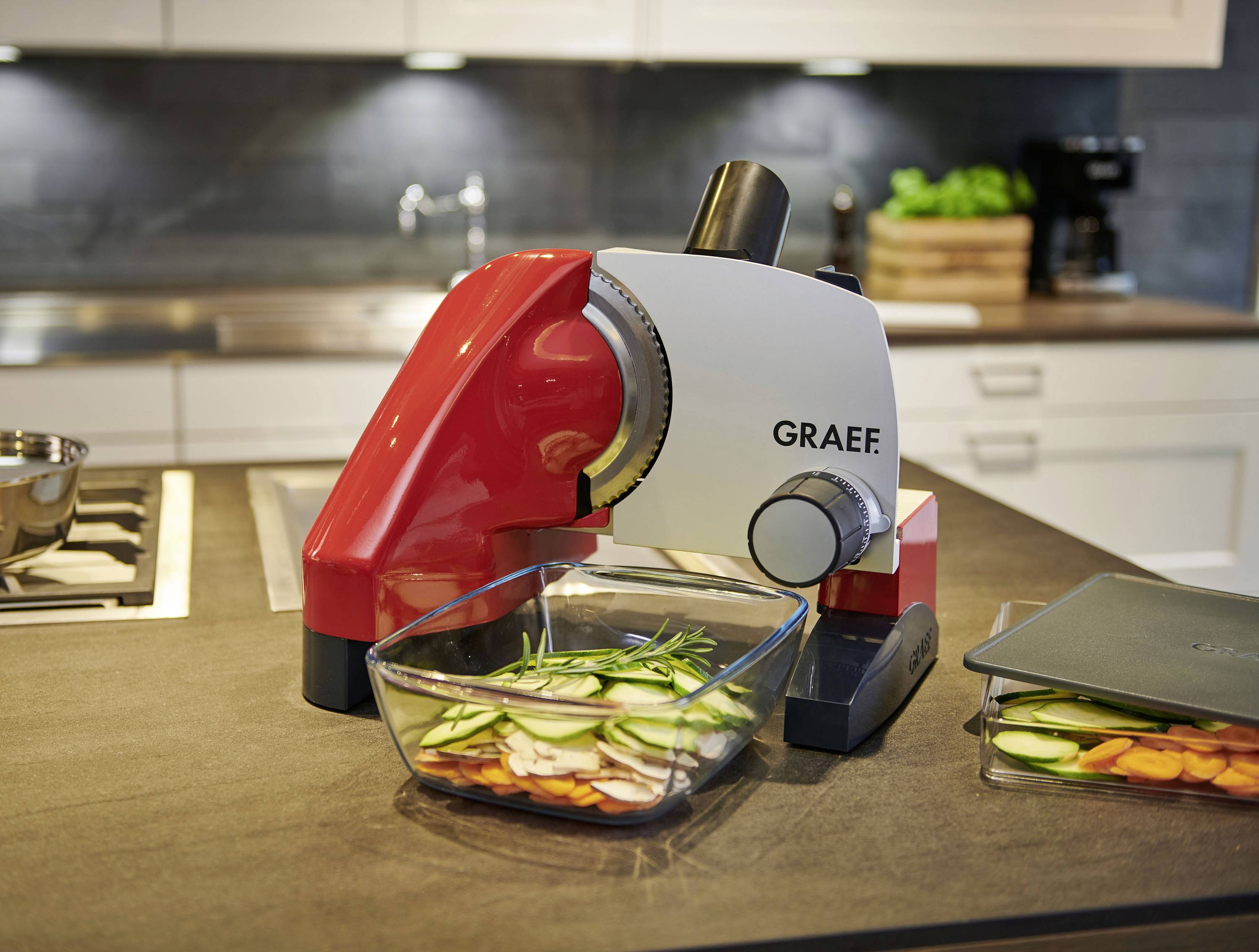 A red and white electric bread slicer sits on a kitchen worktop. Beneath it is a glass bowl filled with chopped vegetables.
