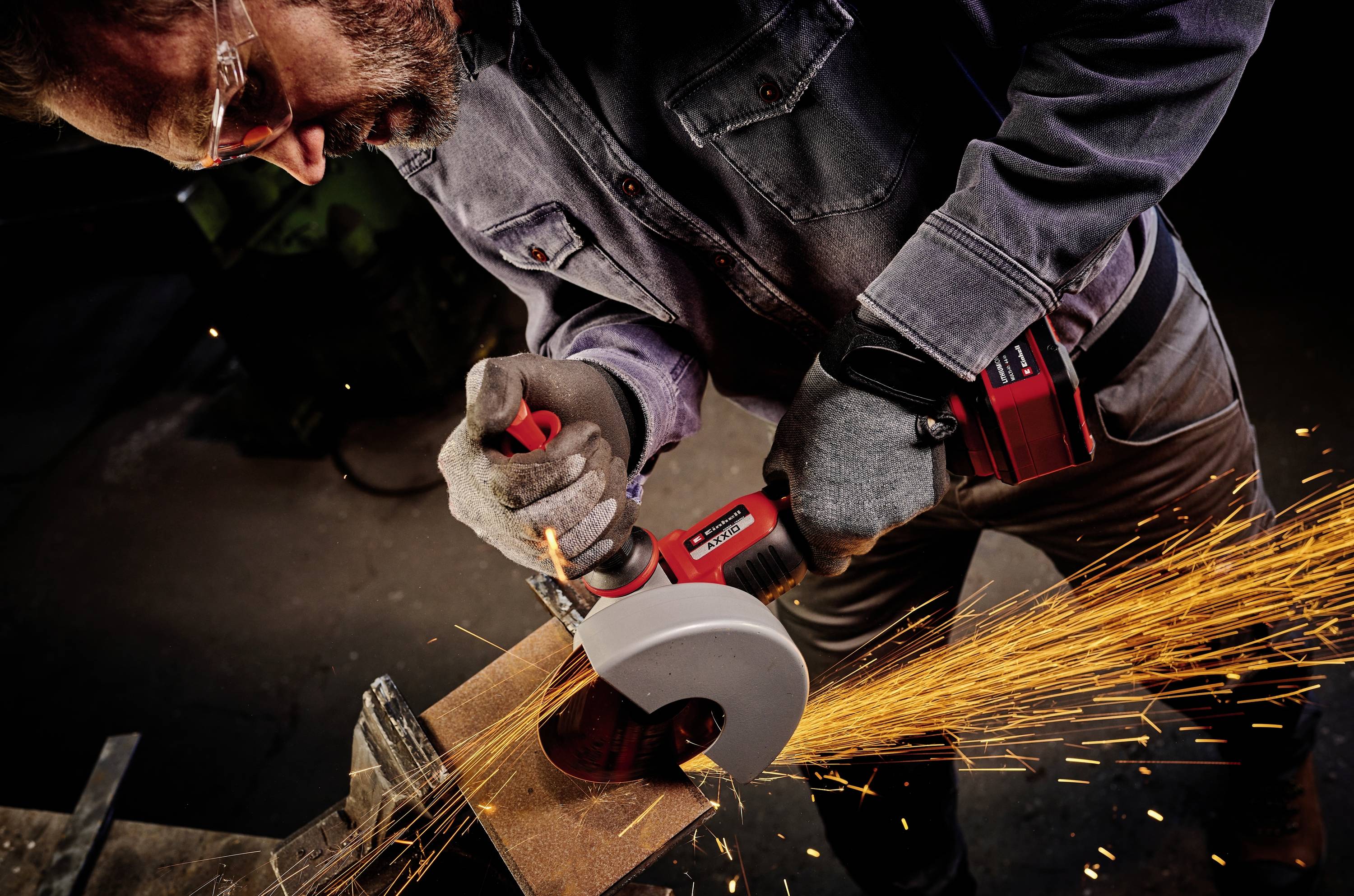 A man is wearing safety glasses and using an angle grinder, with sparks flying as he cuts metal. He is wearing protective clothing.