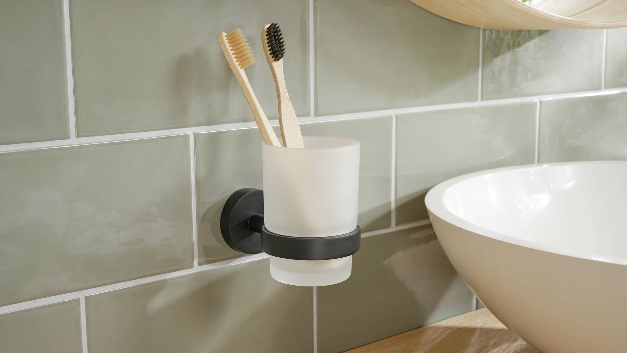 Two wooden toothbrushes in a glass holder beside a sink against green tiles.