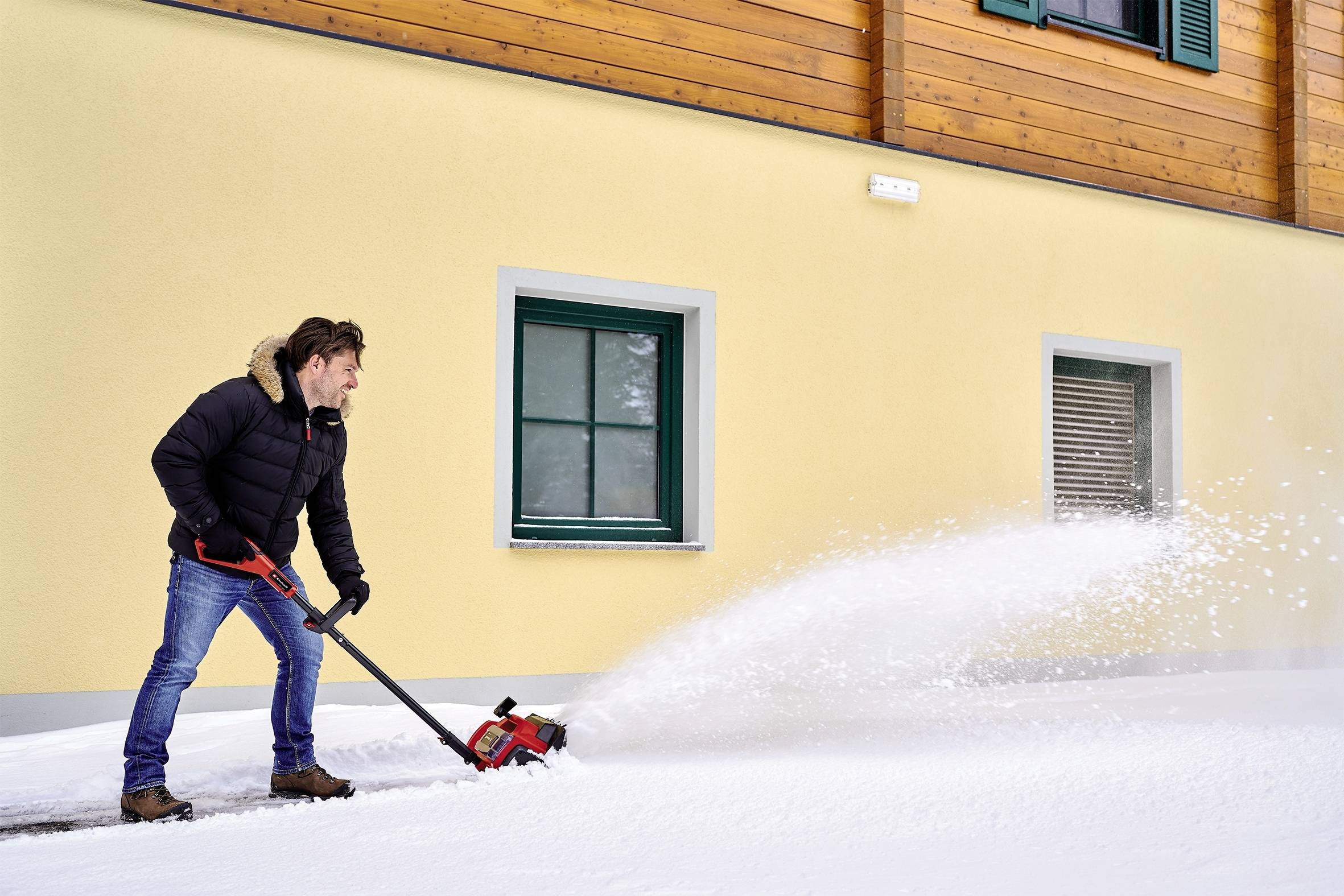 A man is clearing snow in front of a house using a snow blower. He is wearing warm clothing and working on a snow-covered ground.