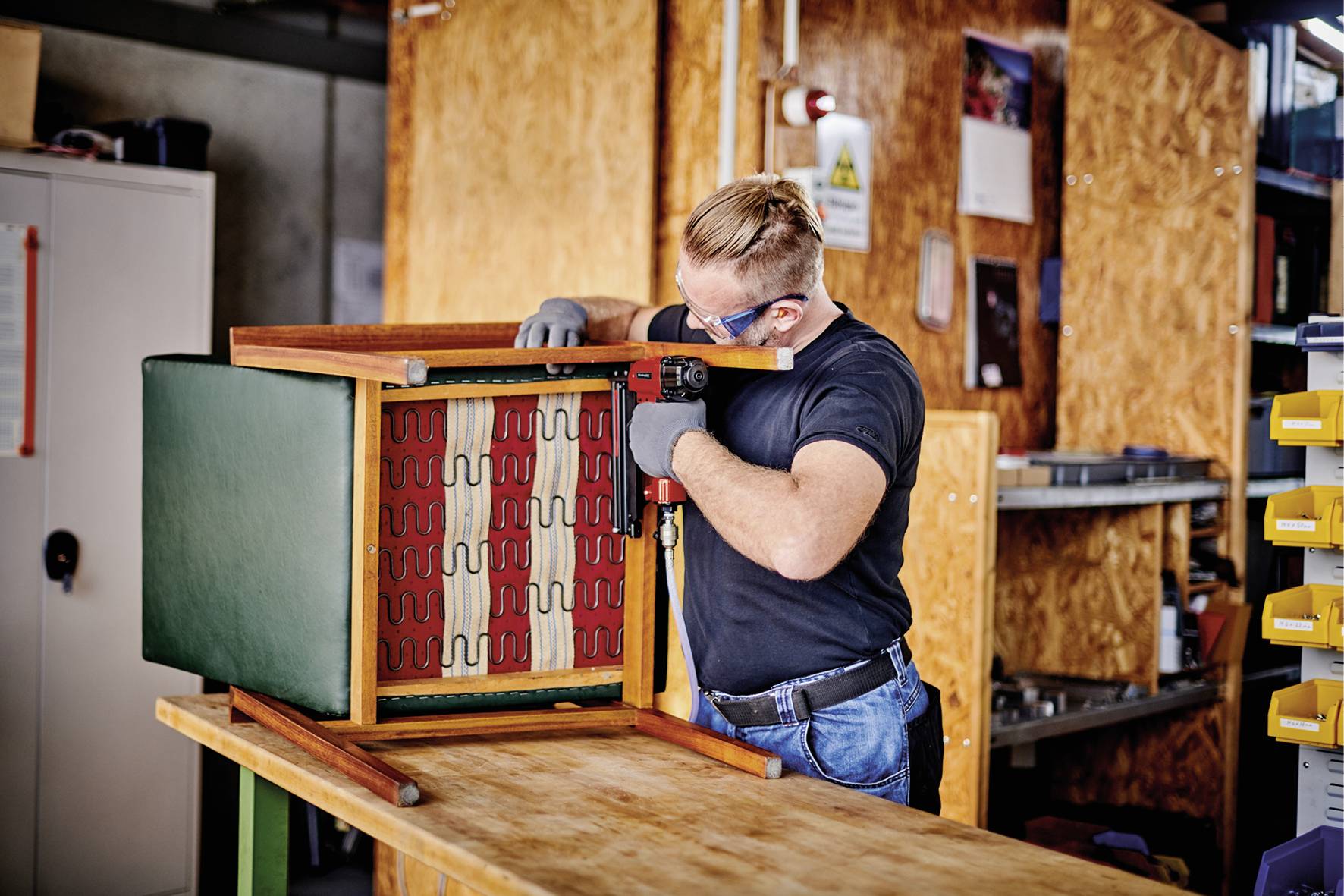 A person is repairing an upturned chair on a table in a workshop. They are wearing safety glasses and gloves.