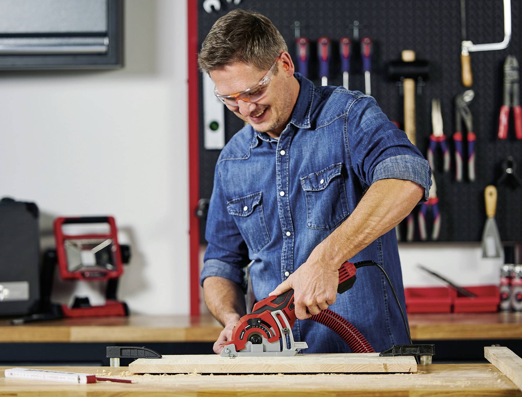A man wearing safety glasses and a denim shirt is working with a circular saw on a piece of wood at a workbench in a workshop.
