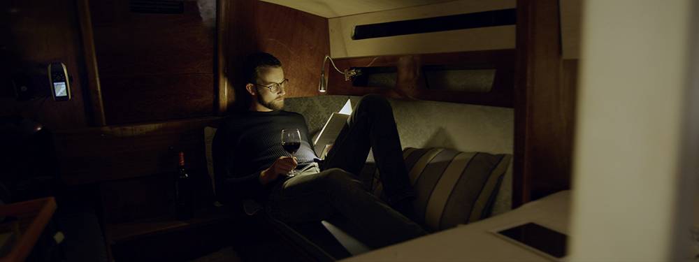 A man sits in a cosy boat, reading a book by lamplight and holding a glass of wine, surrounded by wooden walls.