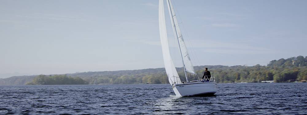 A single sailing boat glides across a large lake in calm weather, with wooded hills visible in the background.