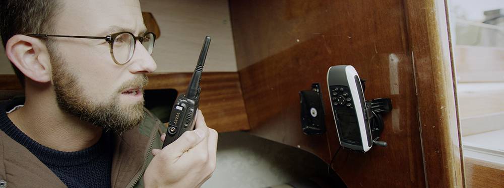 A man is speaking into a two-way radio in a wooden room; another communication device is mounted on the wall.