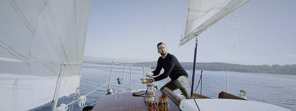 A man is standing on a sailing boat and pulling on a rope. In the background, calm water and a forested coastline can be seen.