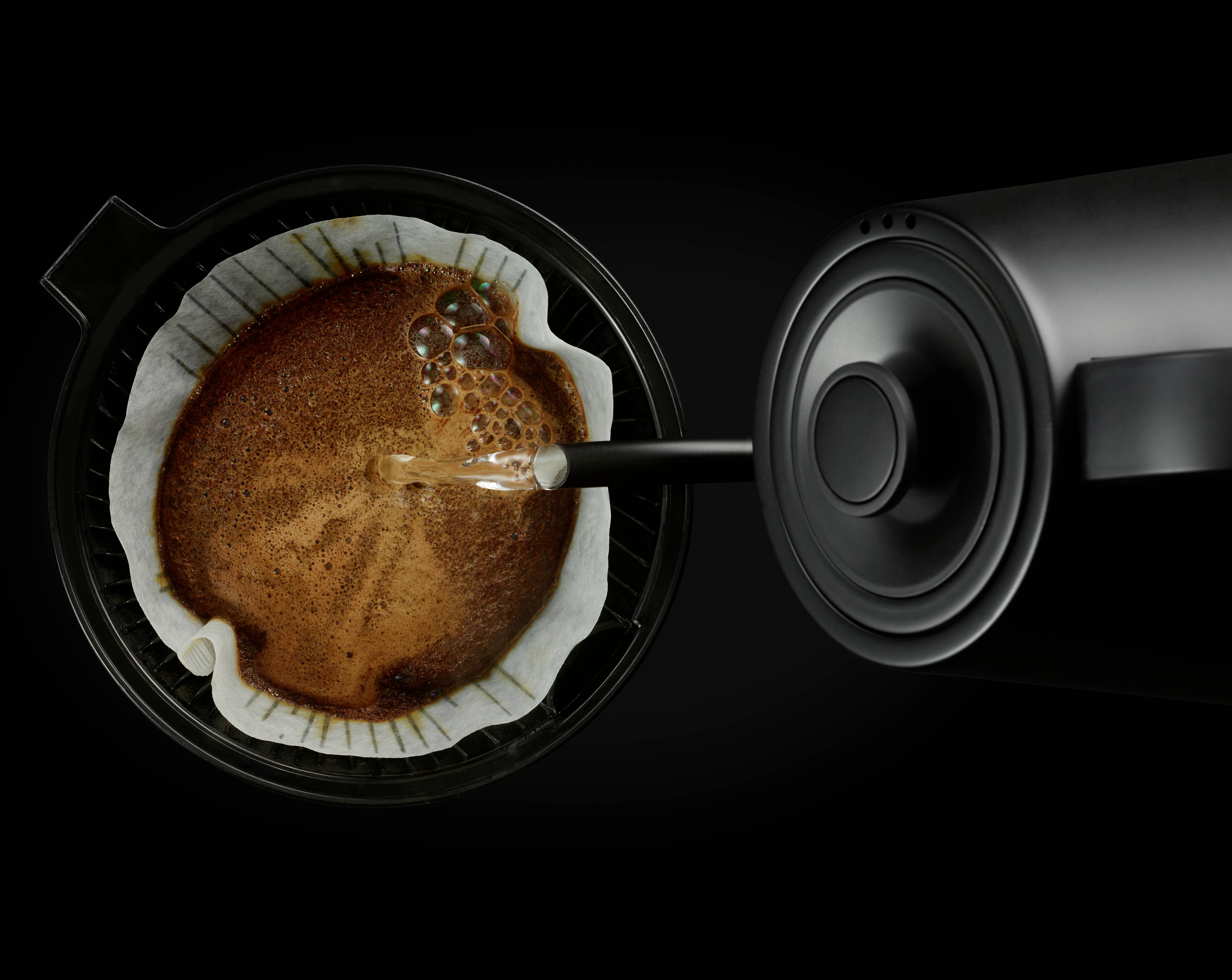 Water is being poured from a black kettle over filter coffee in a coffee machine.