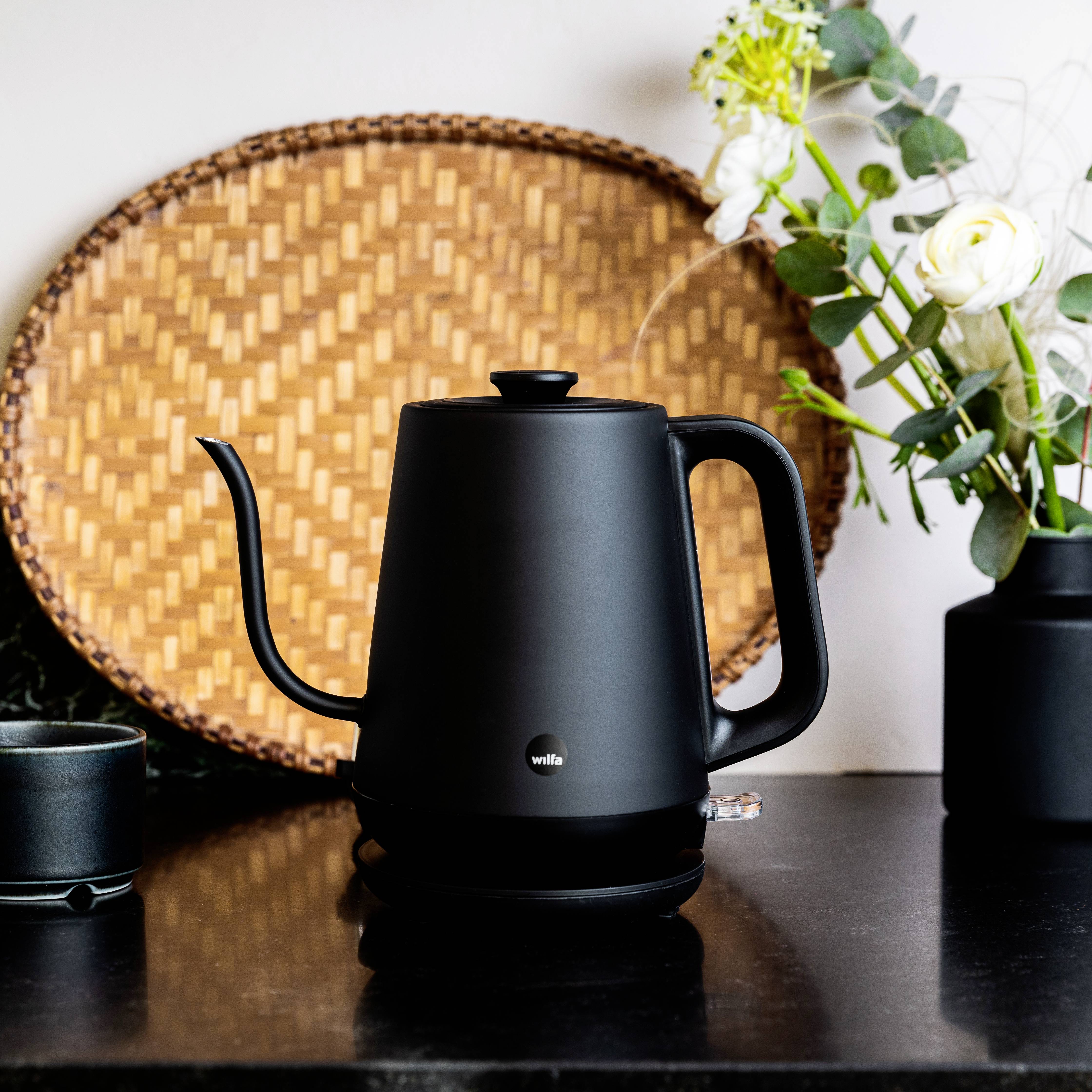 A black kettle is sitting on a kitchen table. Beside it are a black mug and a floral arrangement.