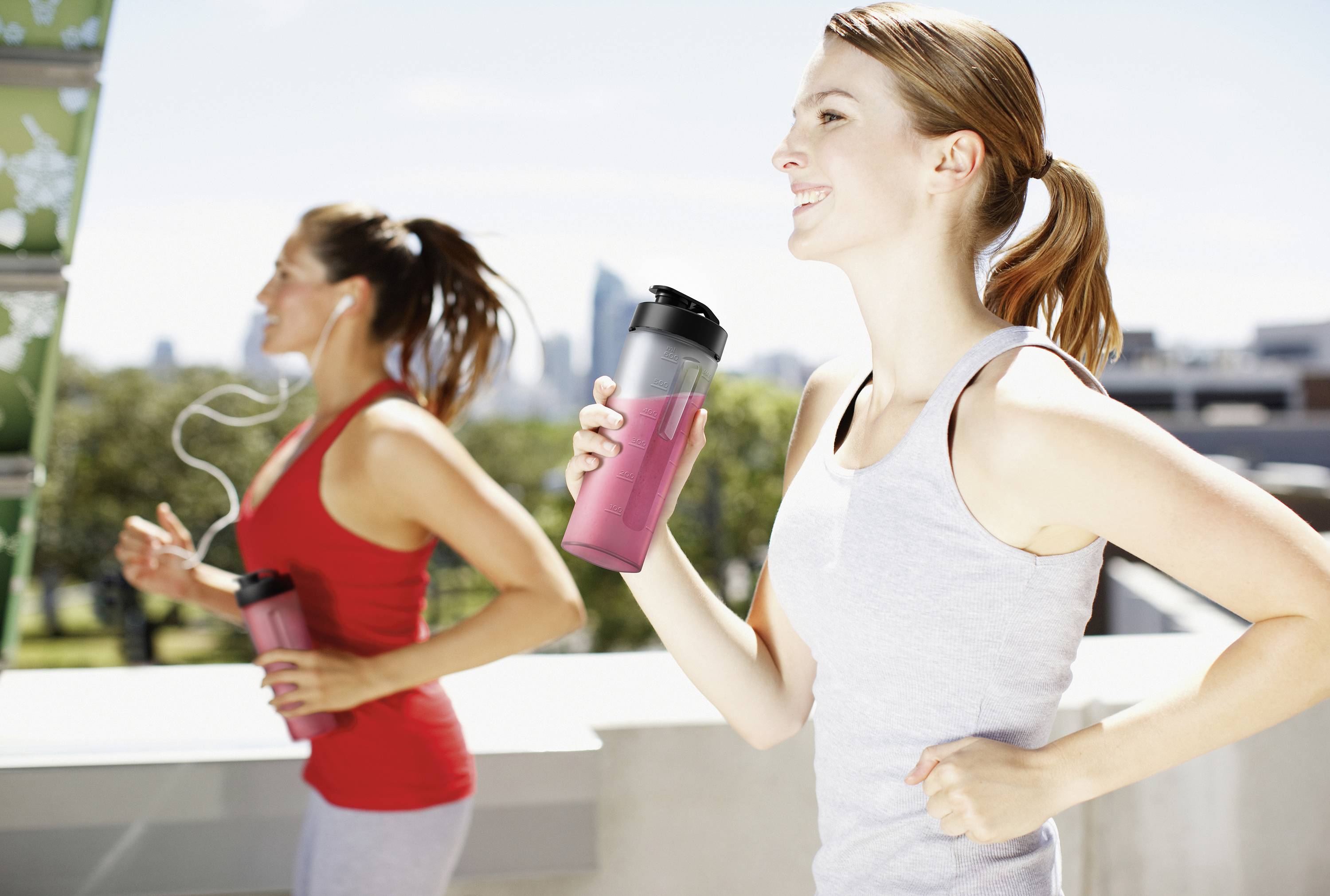 Two women jogging outdoors, holding water bottles, wearing sports clothing, with a blurry city view in the background.
