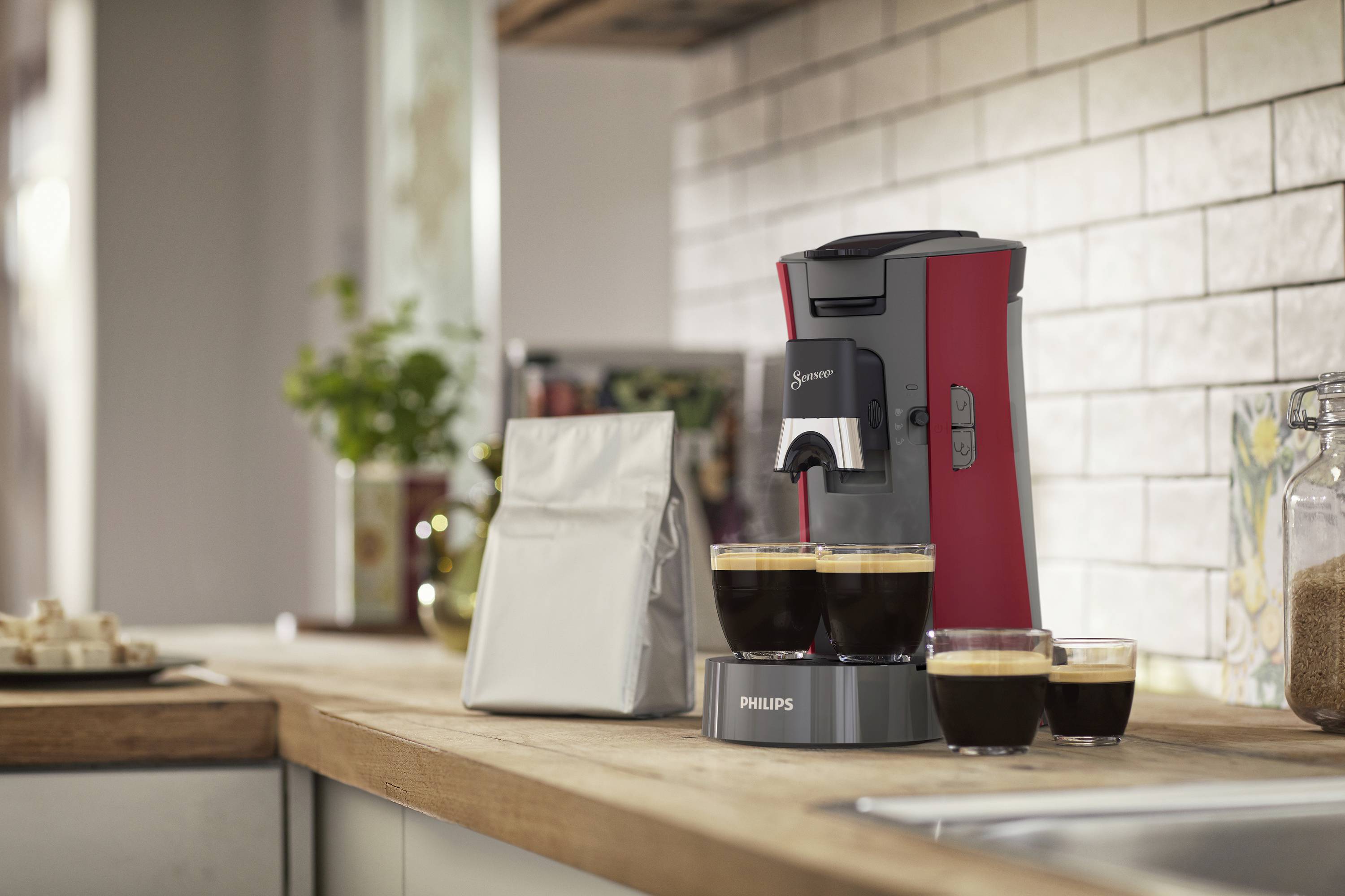 Red coffee machine on a kitchen worktop with two filled cups and a coffee package beside it.