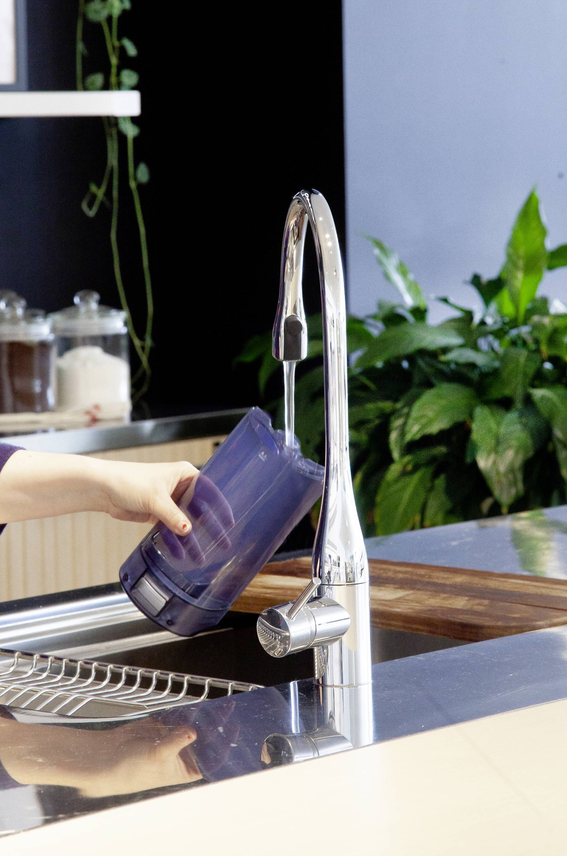 A person fills a dark container at a modern tap in a kitchen with plants in the background.