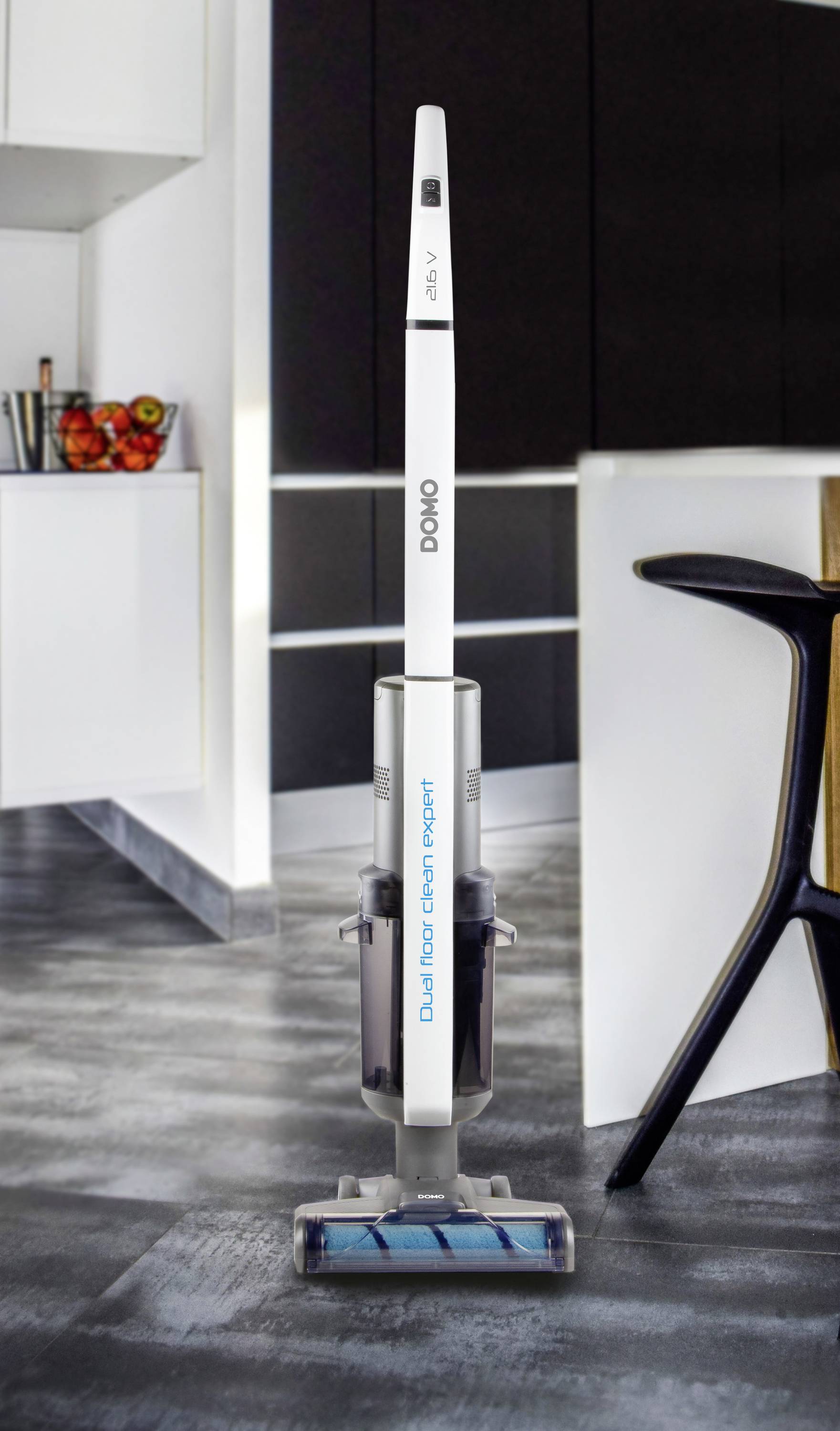 A cordless vacuum cleaner stands on a grey floor in a modern kitchen. Kitchen cabinets and a bowl of fruit are visible in the background.