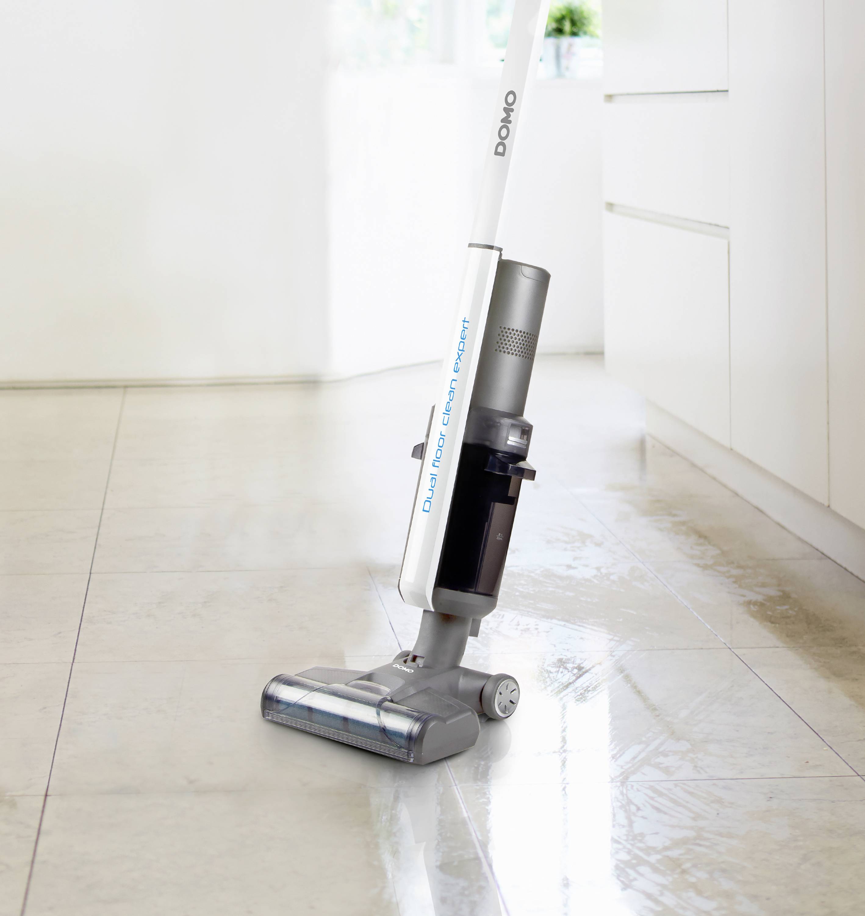 A cordless vacuum cleaner stands on a clean, gleaming kitchen floor. The kitchen is bright with white cupboards.