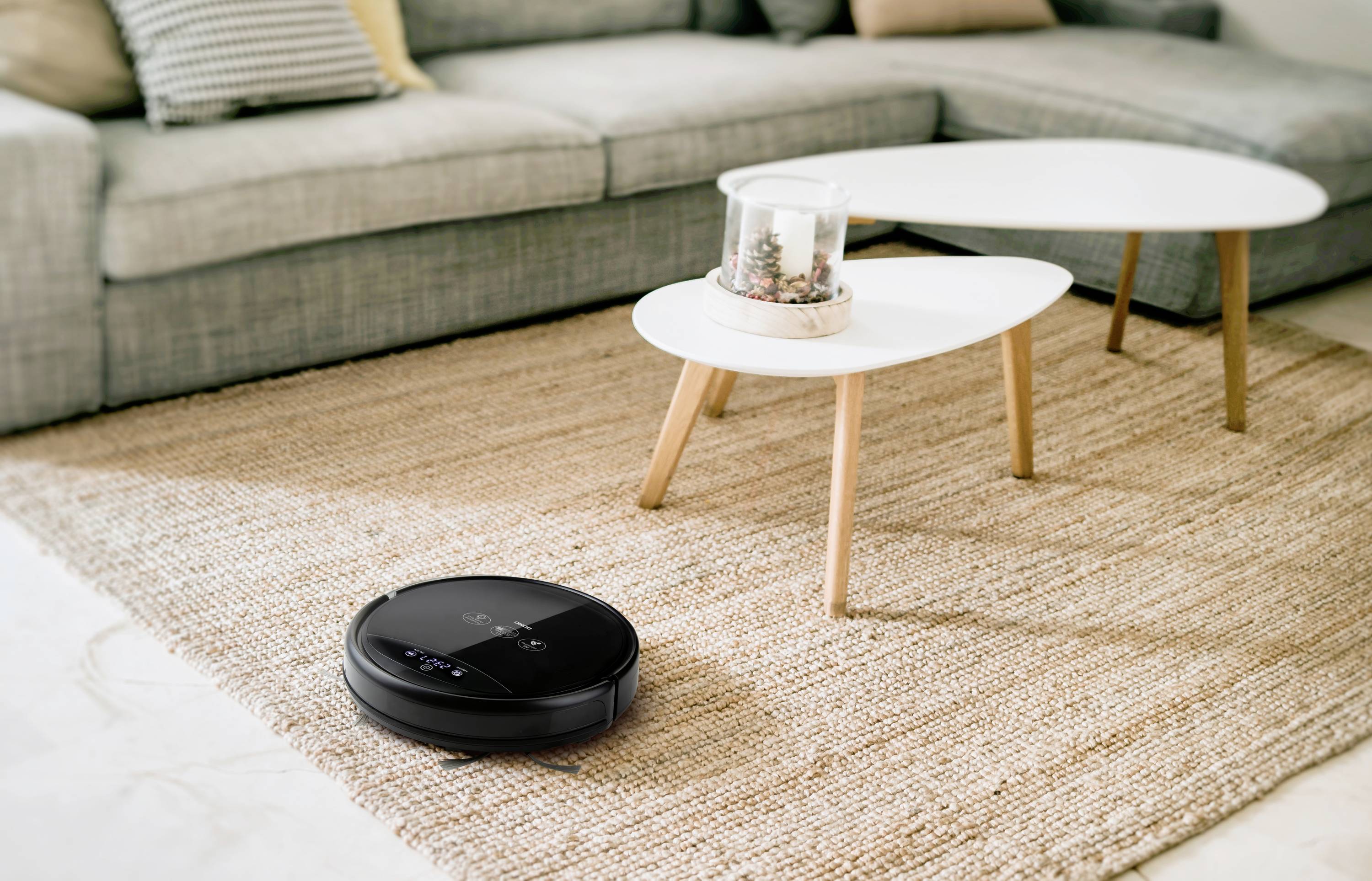 A black robotic vacuum cleaner is cleaning a beige-brown carpet in a living room with a grey sofa and white side tables.