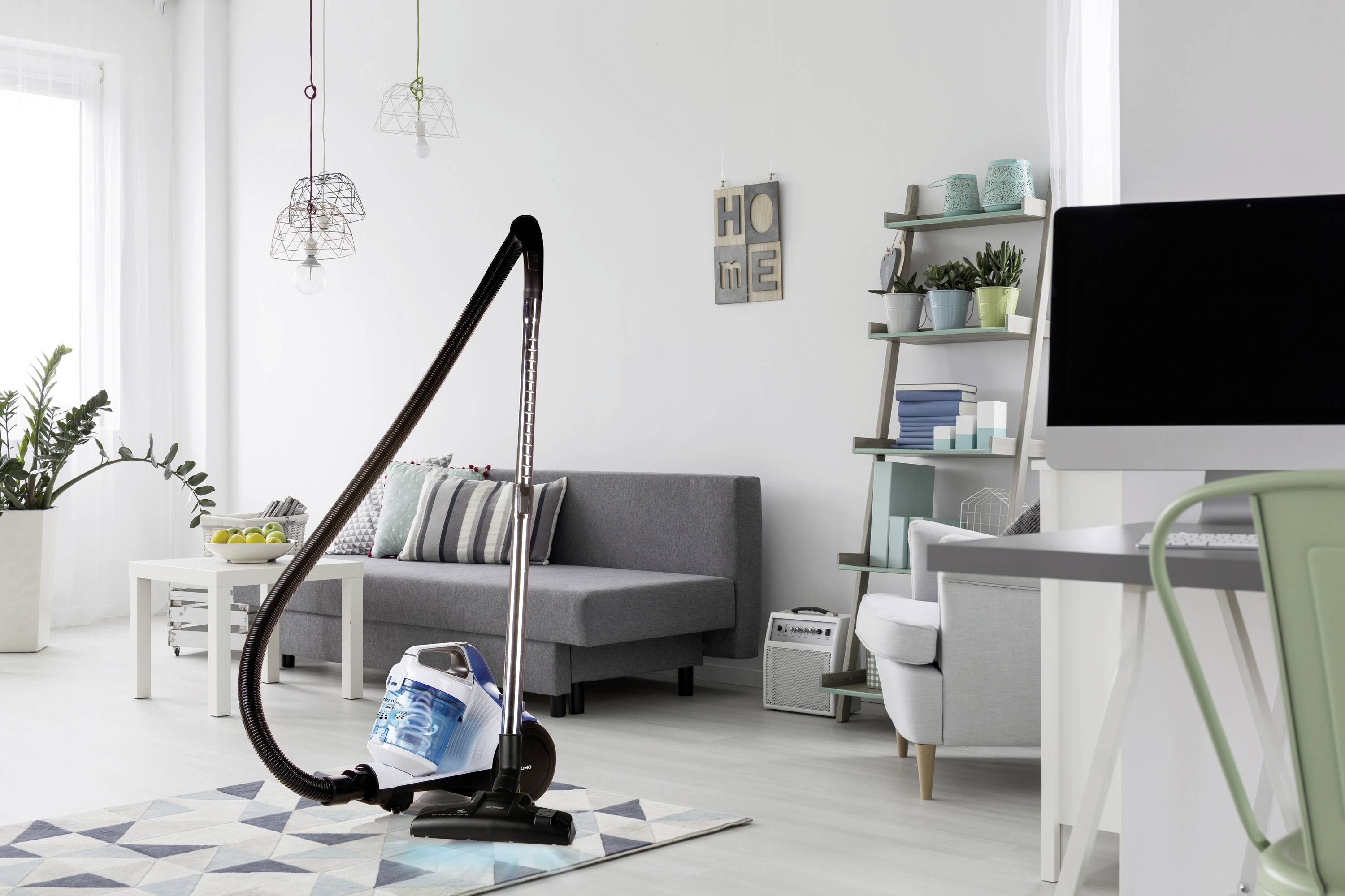A vacuum cleaner stands in a modernly furnished living room. In the background, a grey sofa, bookshelf, and plants can be seen.