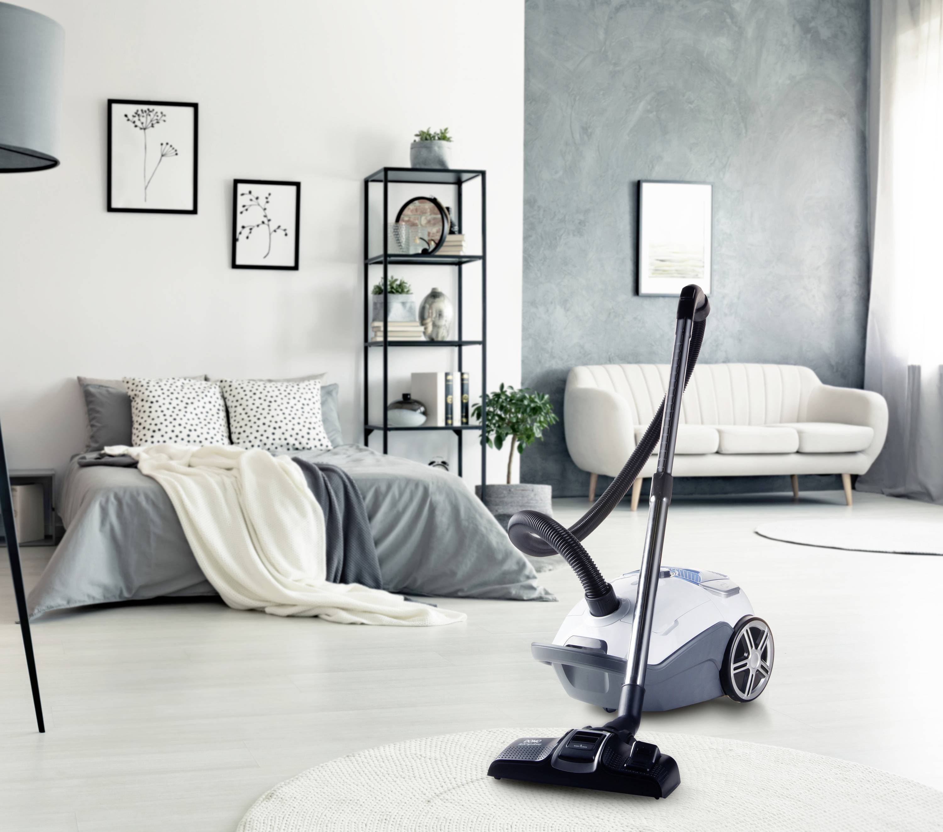A vacuum cleaner stands in a modern, stylish living room with a grey sofa, bed and decorative shelves.