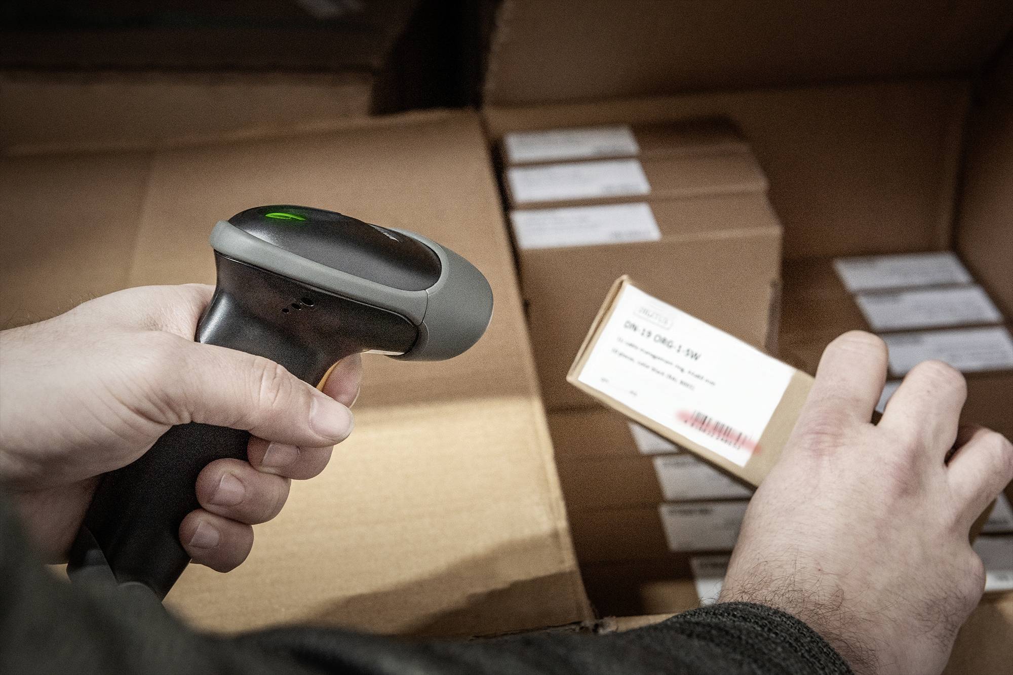 A person is scanning a barcode on a package with a handheld scanner in a warehouse. Additional cardboard boxes are visible in the background.