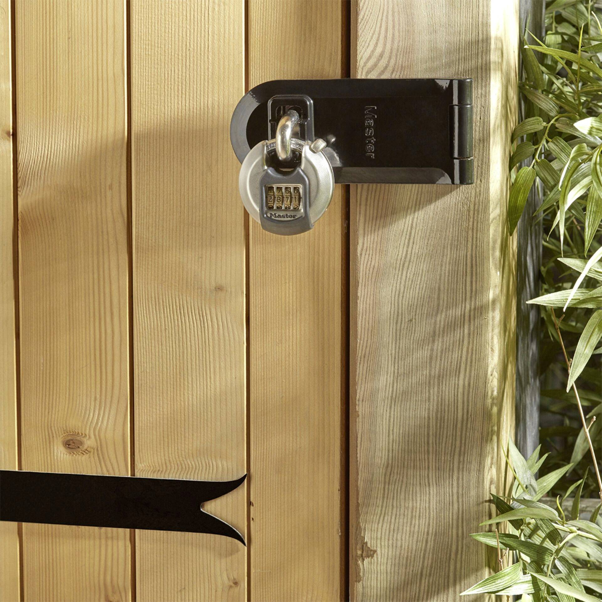 A wooden gate with a silver padlock attached. The gate is closed, and plants are visible in the foreground.