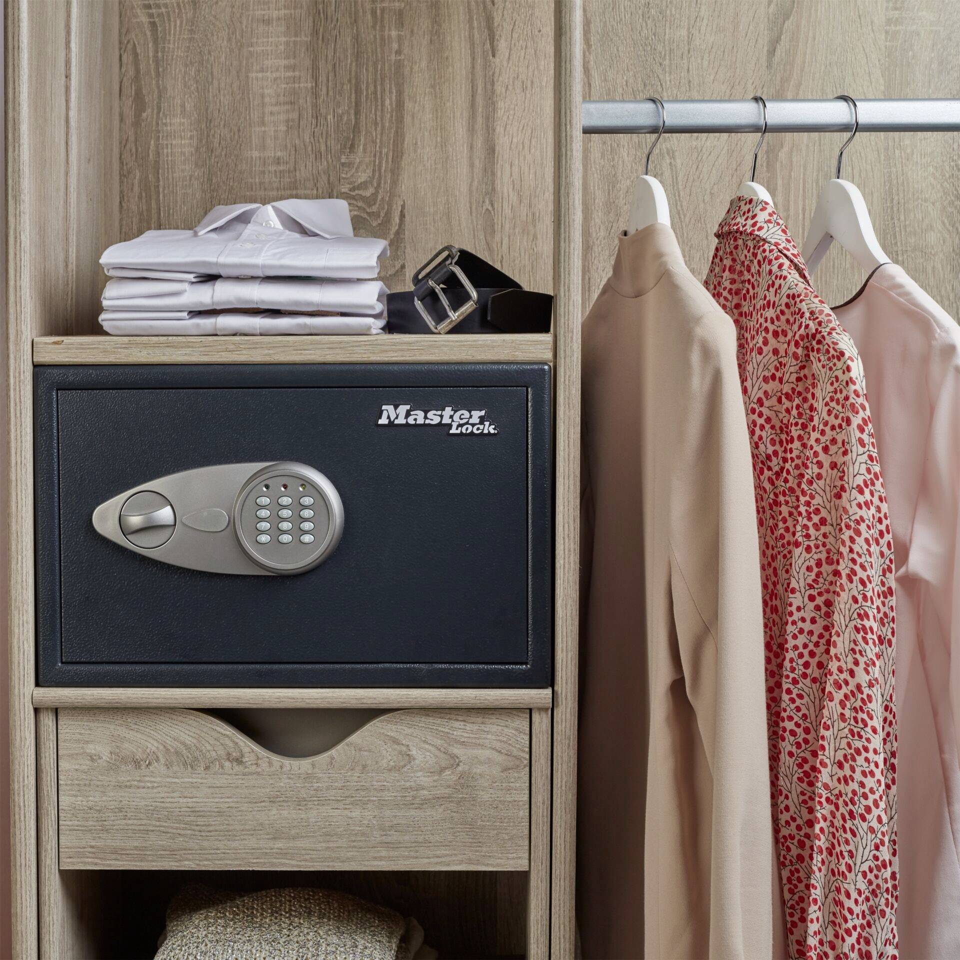 A black safe with a number lock is situated in a wooden wardrobe. To the right, shirts and a patterned dress are hanging.