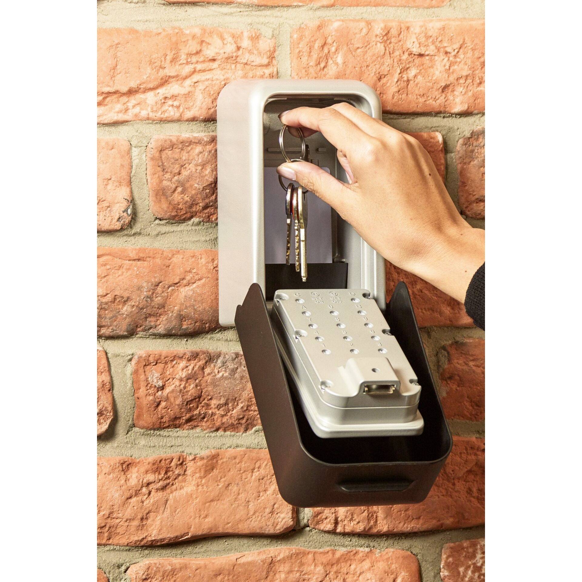 A hand takes a bunch of keys from an open key safe mounted on a brick wall. The safe is rectangular and dark grey.