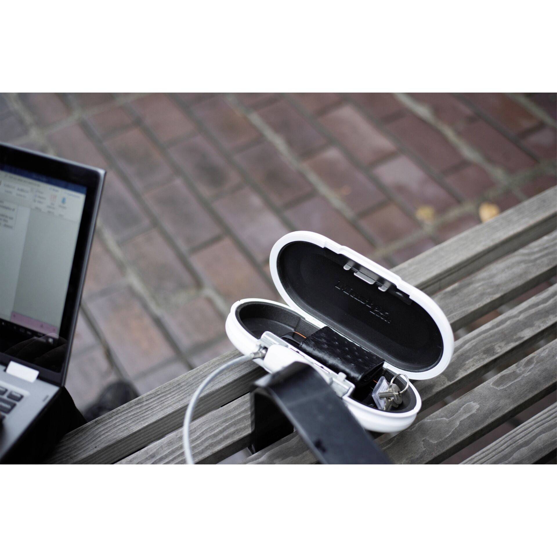 A laptop is charging a pair of headphones in a white box on a wooden bench. Paving stones are visible in the background.