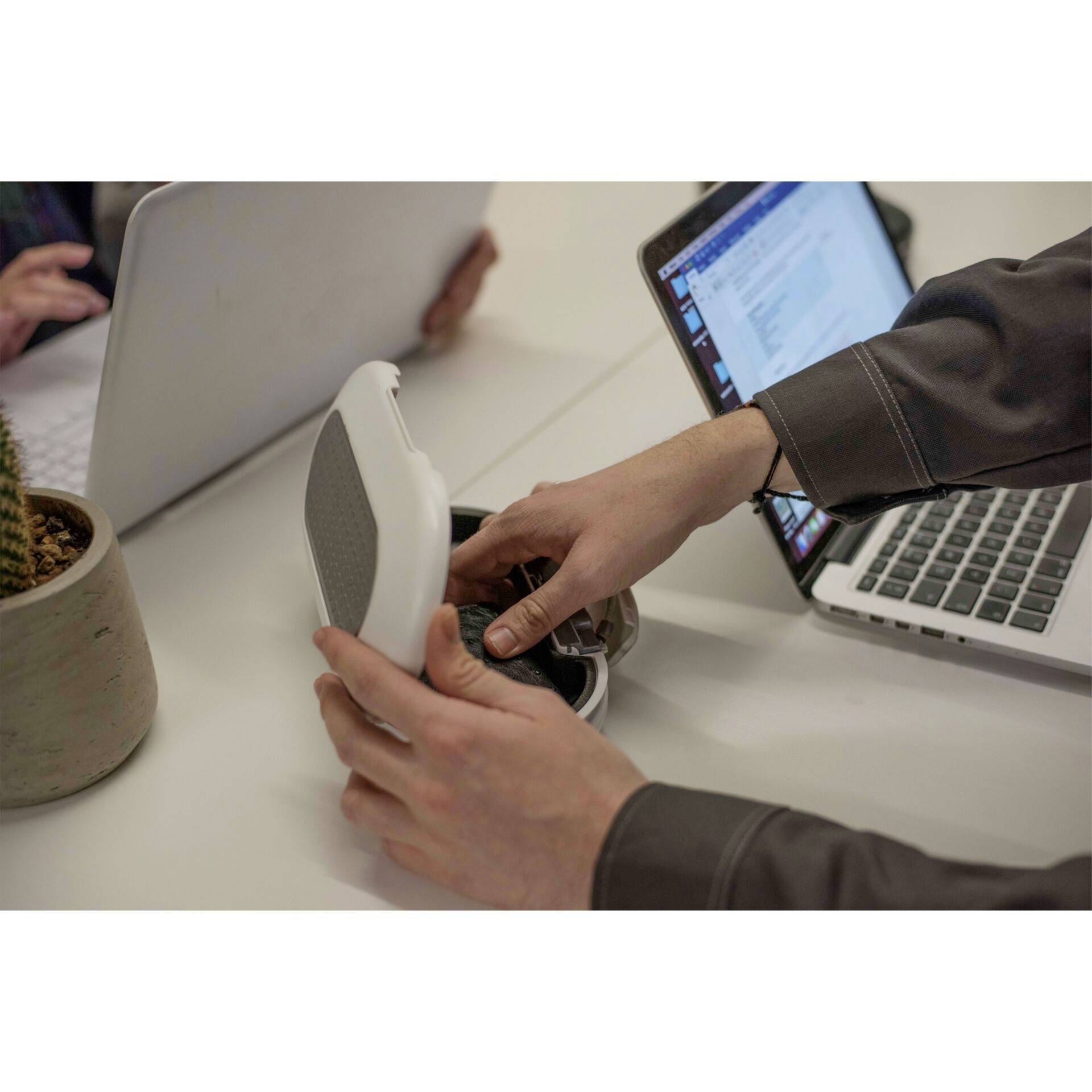 Two people are working on laptops at a table. One person is holding a white round device, possibly a speaker, in their hand.