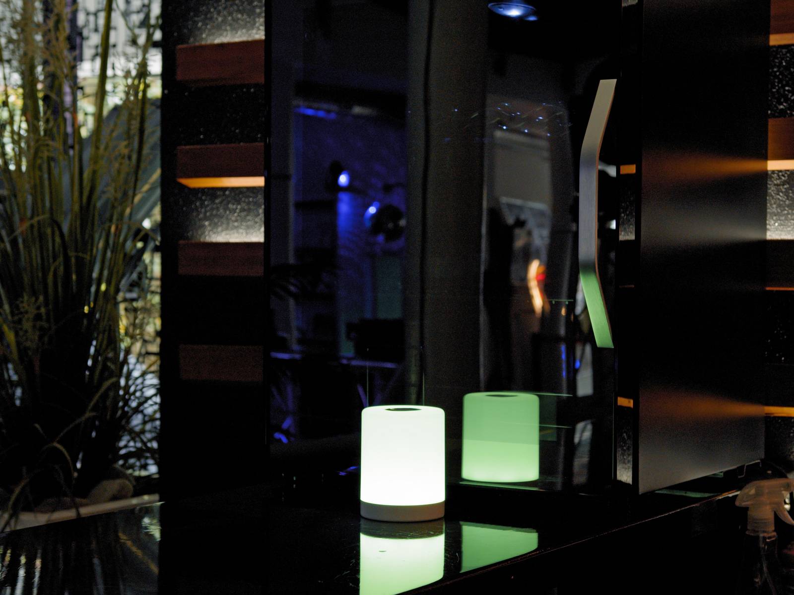 A small, green luminous cube sits on a dark table, surrounded by reflective surfaces in a dimly lit room.