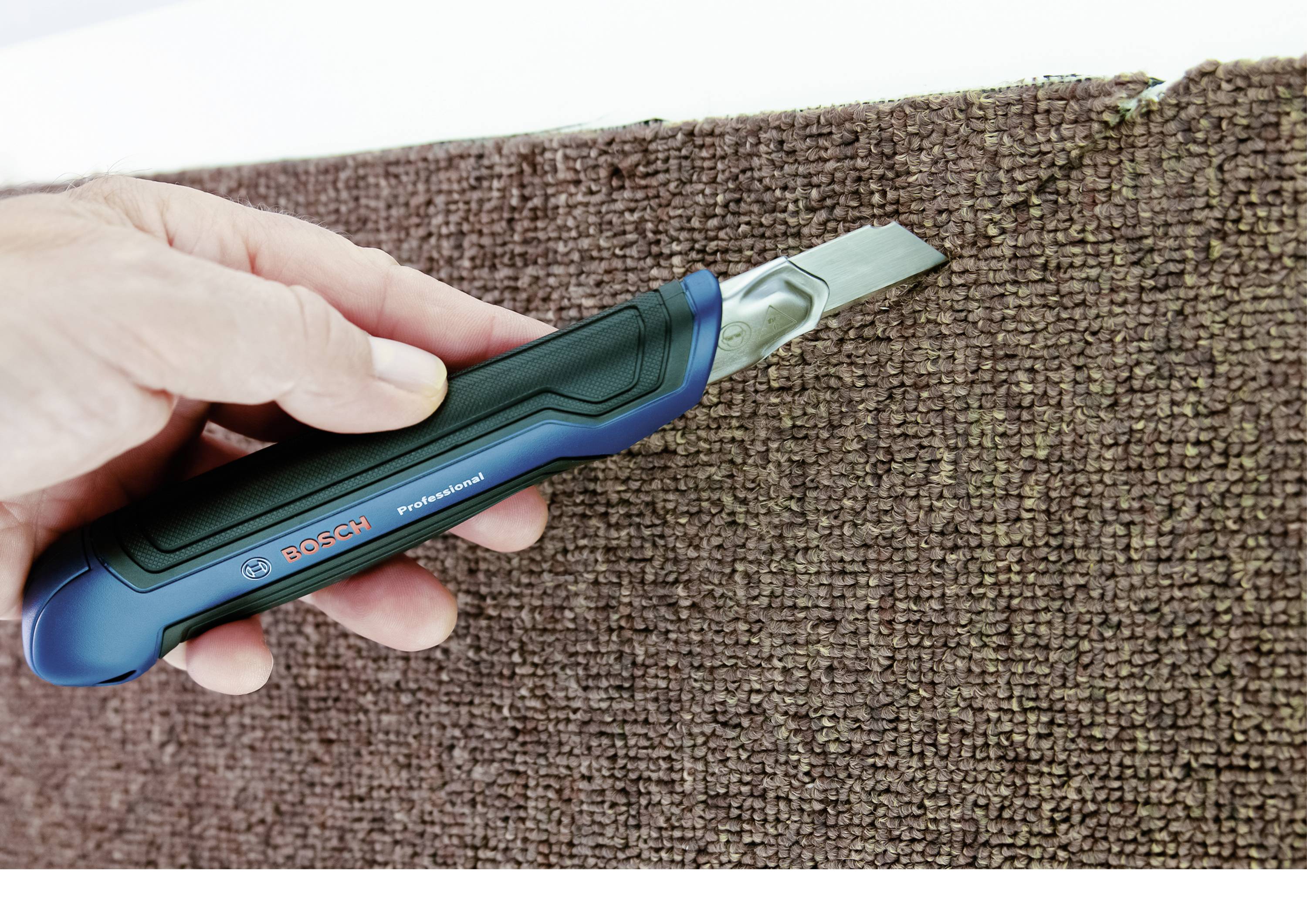 A hand is cutting a brown carpet with a blue carpet knife.