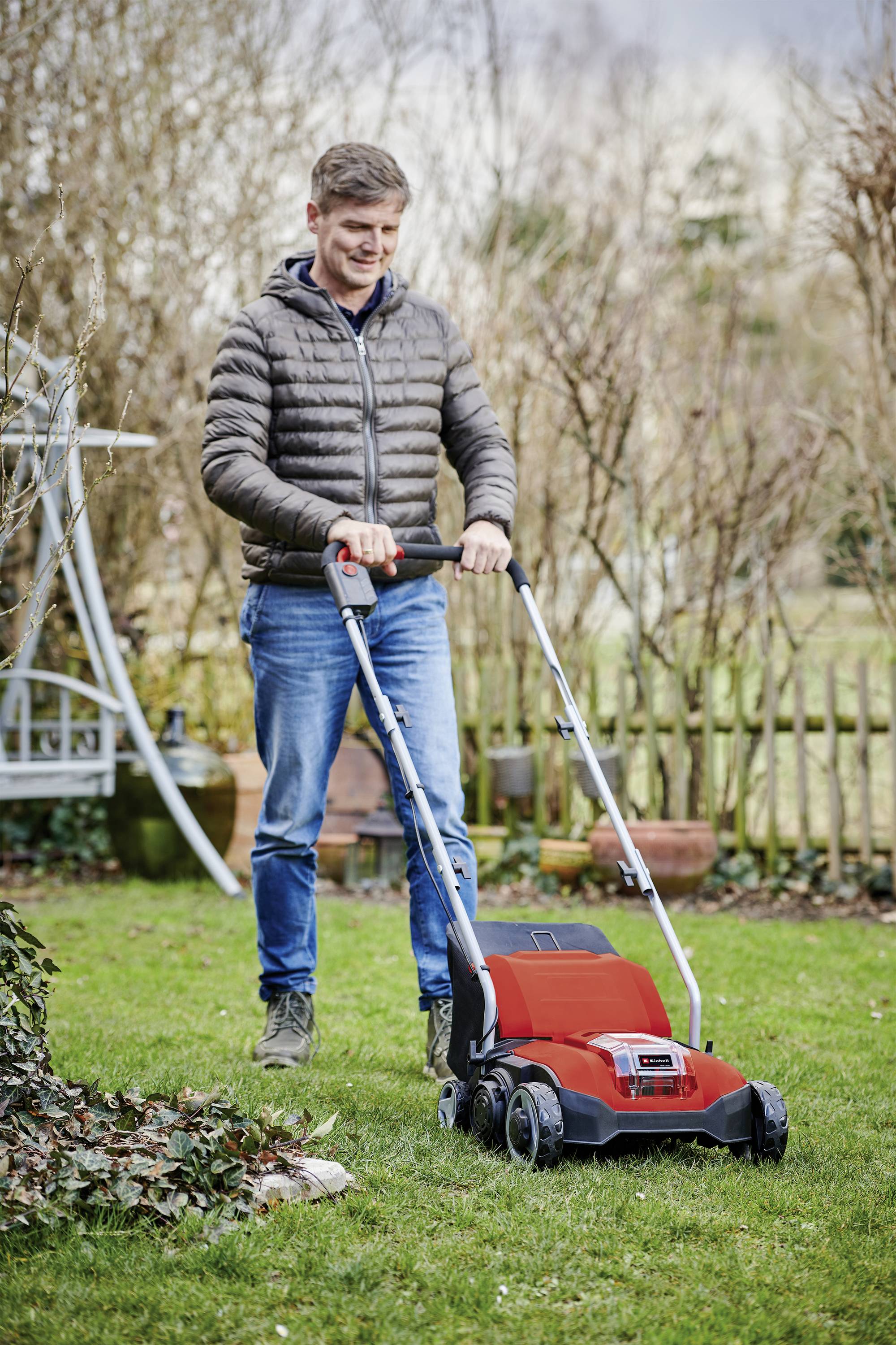 A man in casual clothing is pushing a red lawnmower across a green lawn in a garden with a wooden fence and bare shrubs.