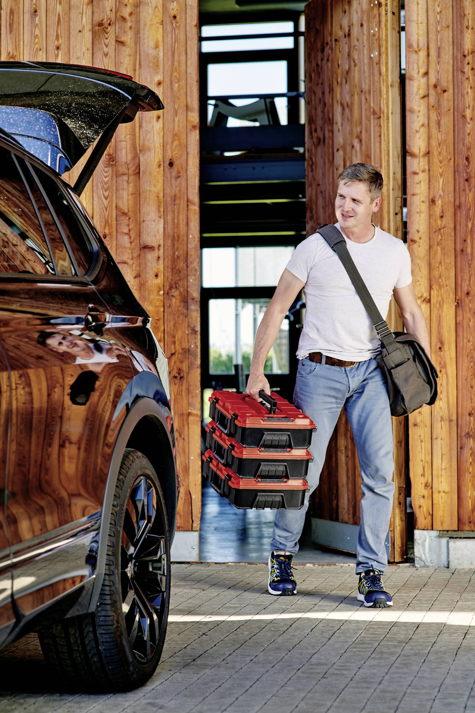 A man in jeans and a T-shirt is holding two red toolboxes and walking towards a car with an open boot. A building entrance is visible in the background.