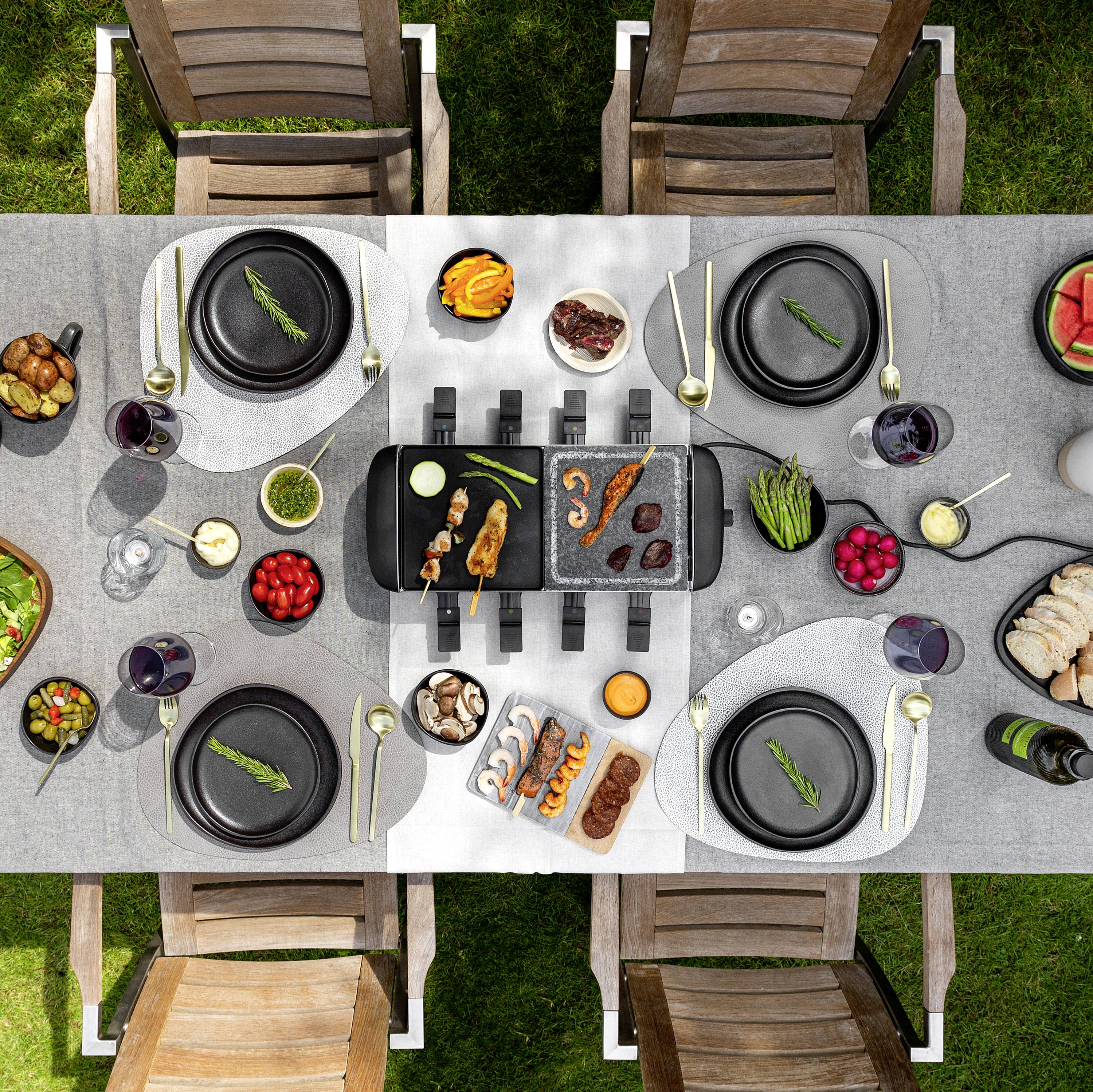 An outdoor dining table is set with an electric barbecue in the centre. Various dishes, salads, drinks and crockery can be seen.