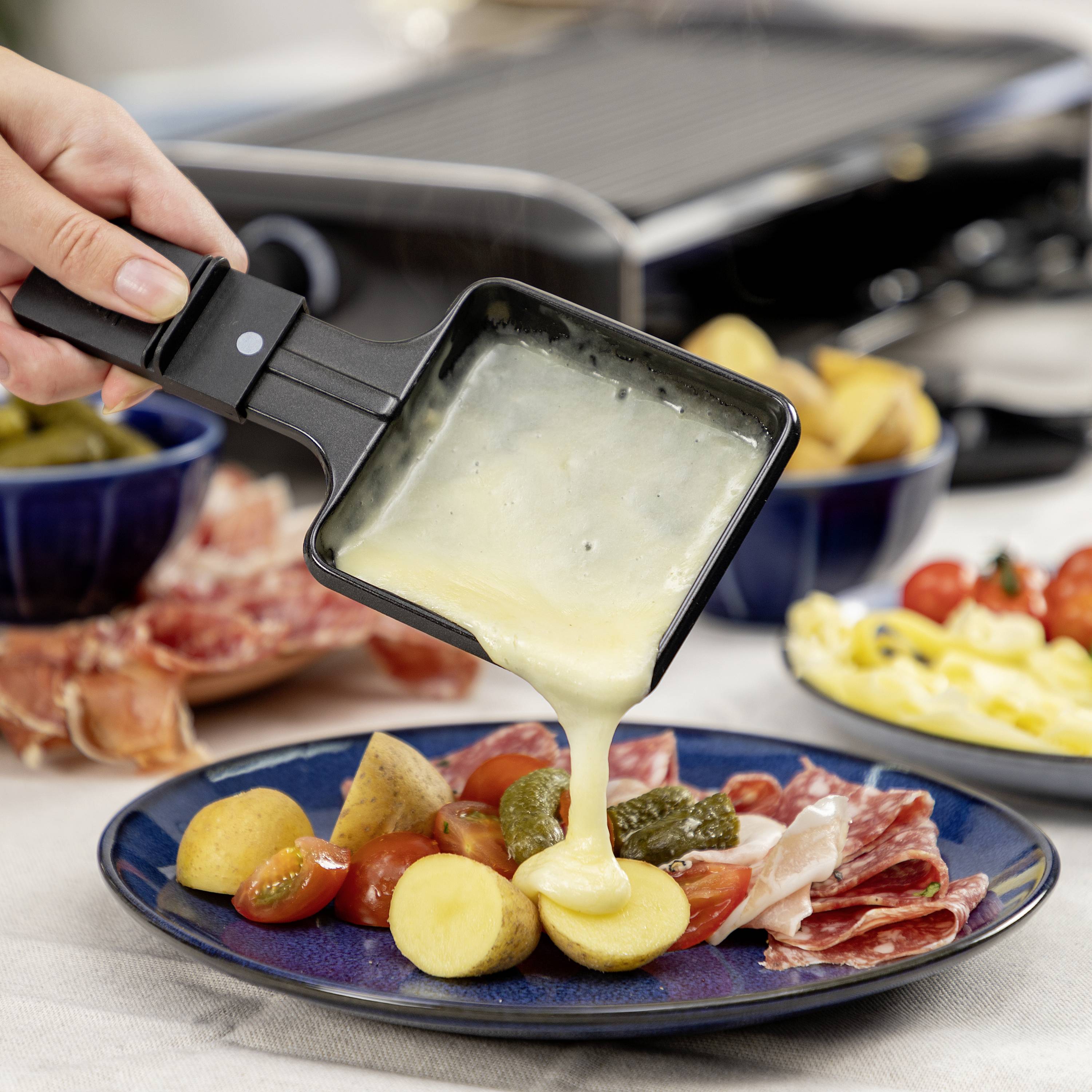 A person is pouring melted cheese over a raclette with potatoes, gherkins and sausage. A grill is visible in the background.