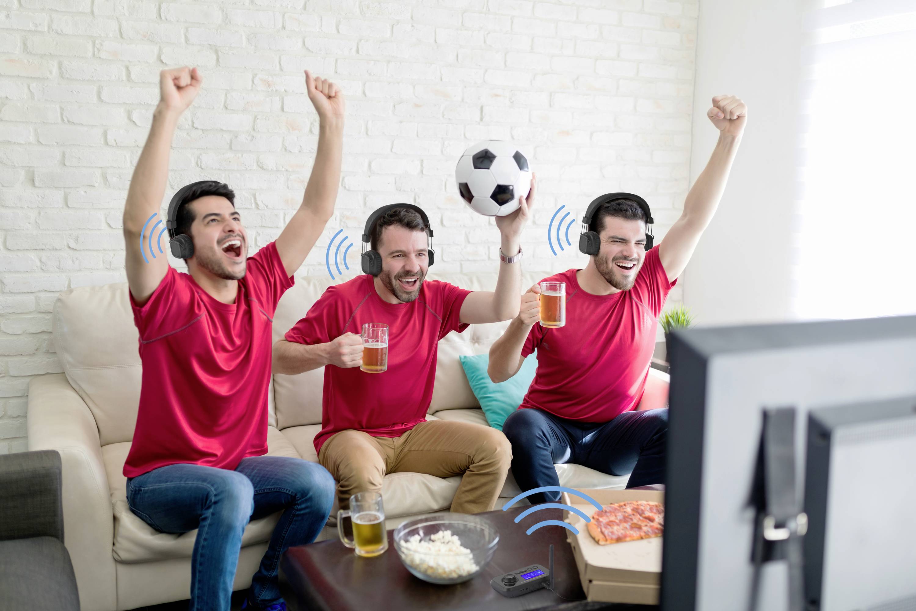 Three men in red jerseys are cheering in front of a television, holding drinks and a football while watching a match.