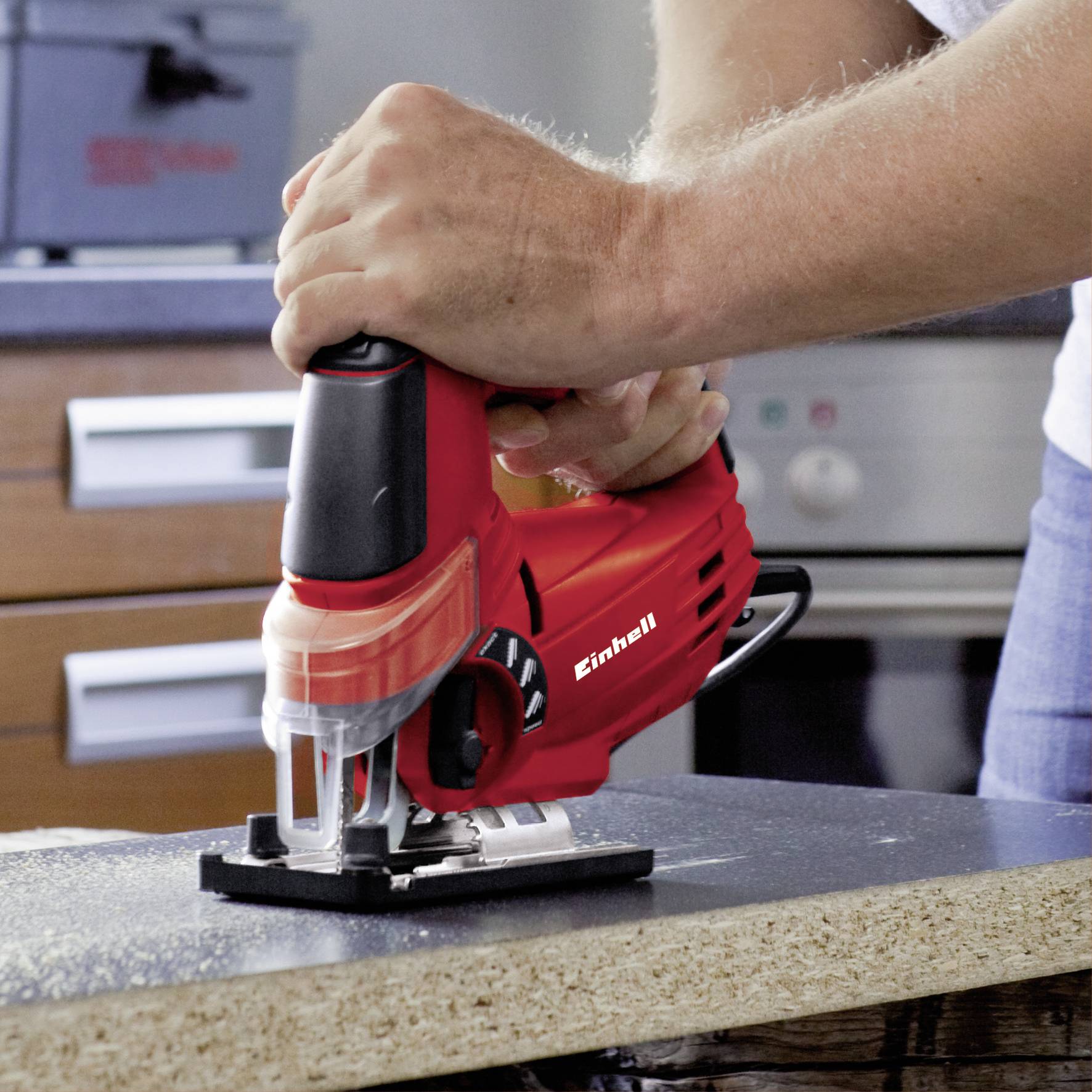 A person is using a red electric jigsaw to cut a piece of wood in a workshop or kitchen.