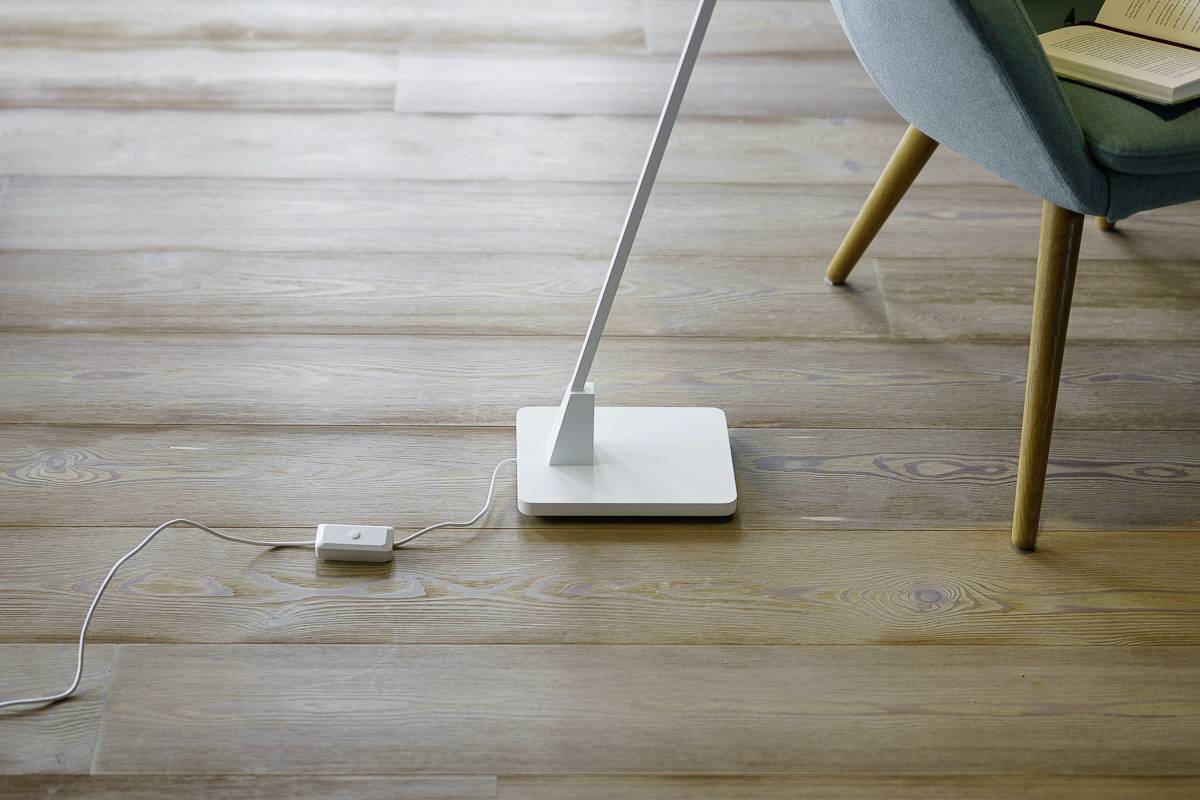 A modern, white standard lamp stands on a light wooden floor. A remote control lies on the ground beside it. Part of a blue chair is visible.
