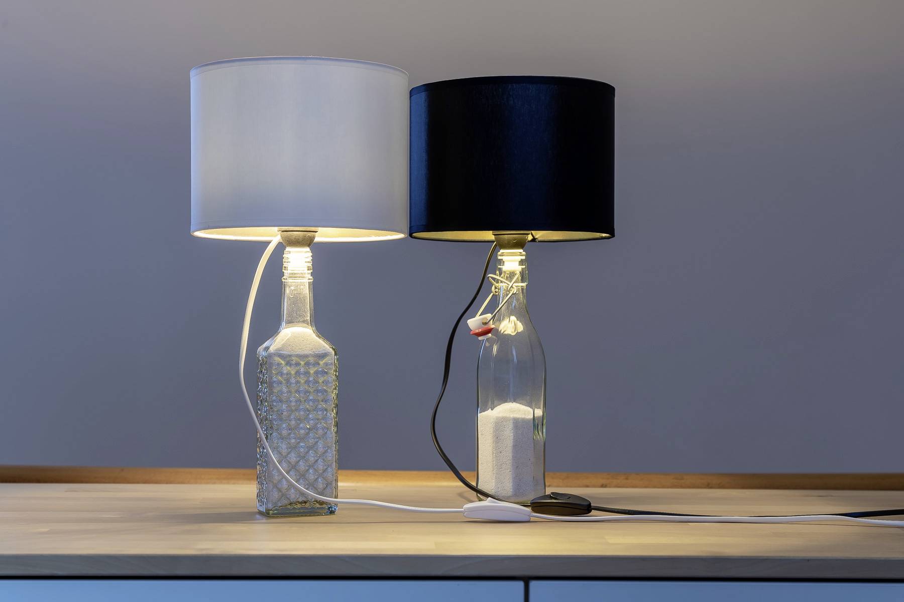 Two table lamps on a wooden cabinet, one with a white shade and the other with a black shade, both made from bottles.