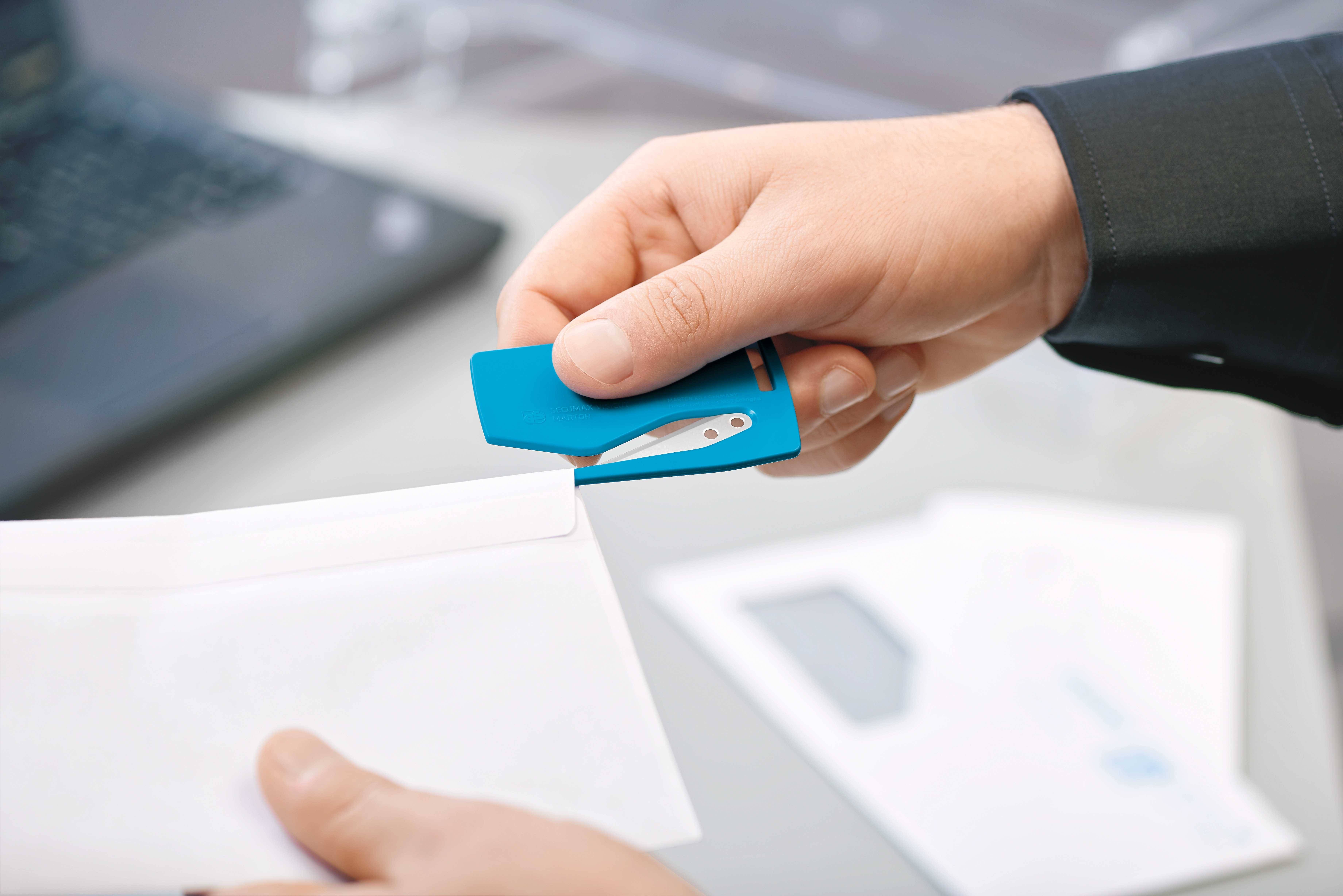 A person is holding a blue paperclip and attaching it to a white envelope. A laptop is visible in the background.