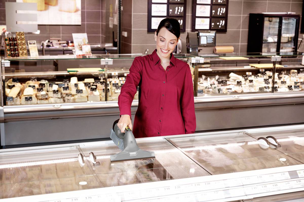 A woman is cleaning the glass surface of a refrigerated display counter with a handheld vacuum cleaner. Various types of cheese can be seen in the background.