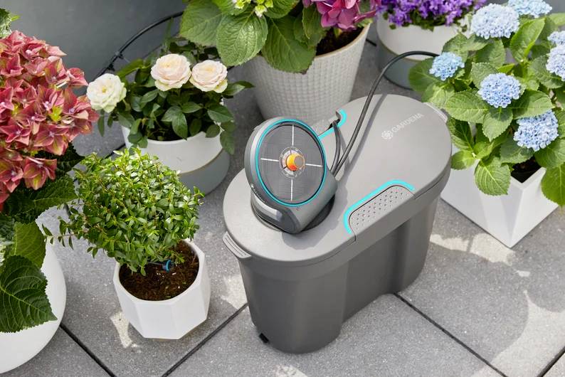A garden irrigation device is placed among potted flowering plants on a tiled patio, indicating its use for automatic plant watering.