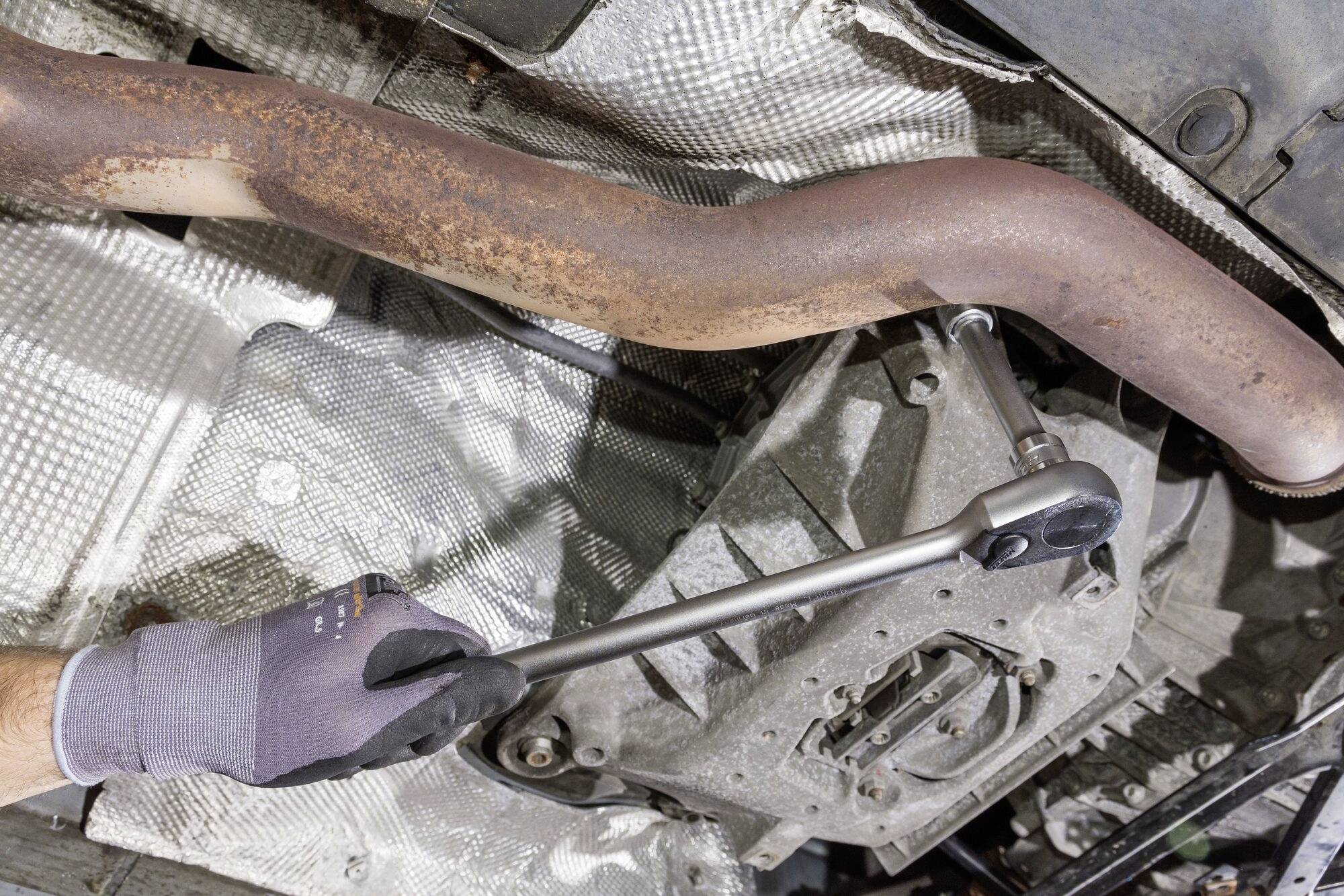 A mechanic's hand wearing a glove uses a ratchet to loosen a screw on the exhaust system underneath a vehicle.