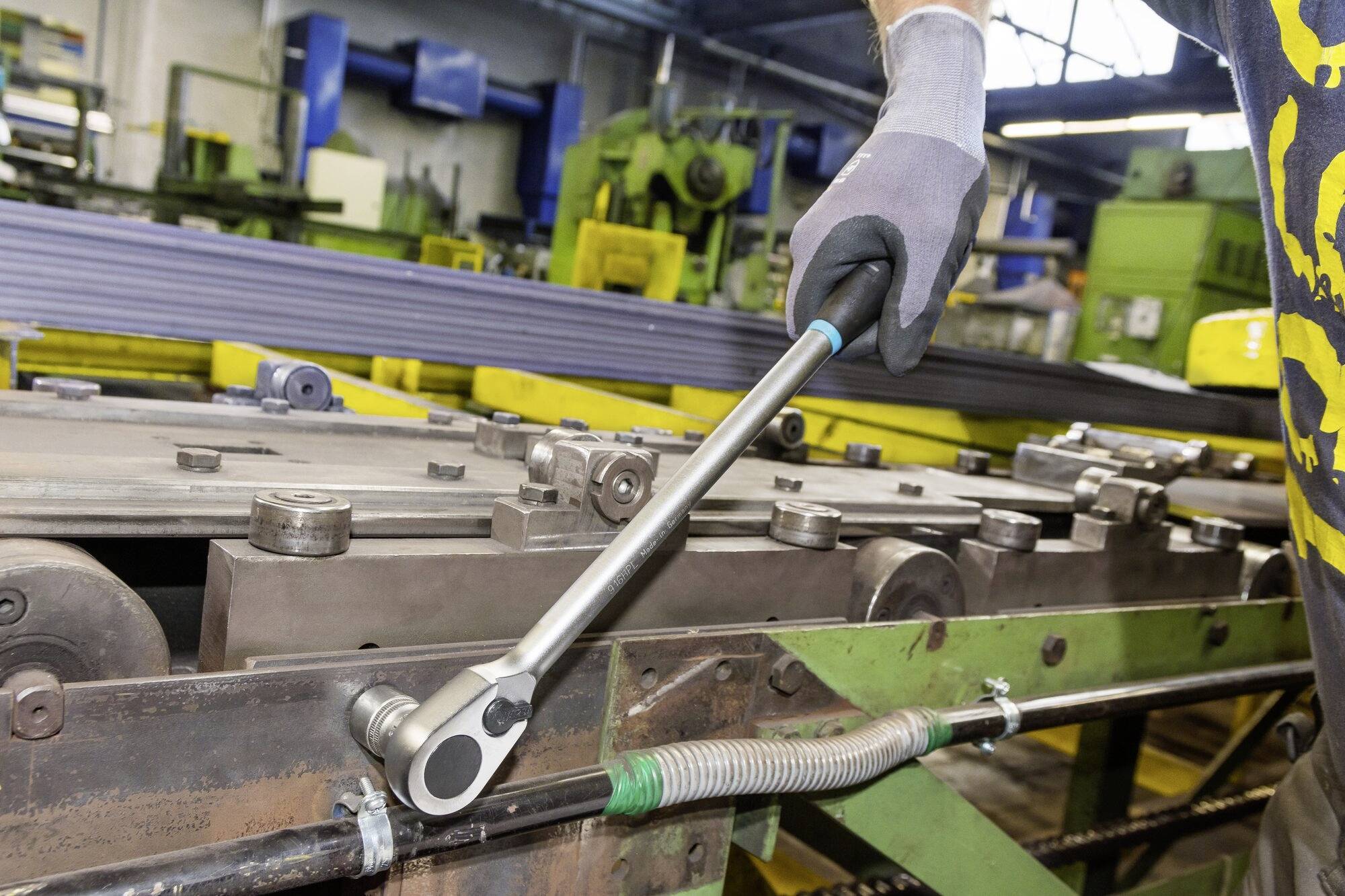 A worker is using a torque wrench in a factory environment to maintain machinery. Green machines are visible in the background.