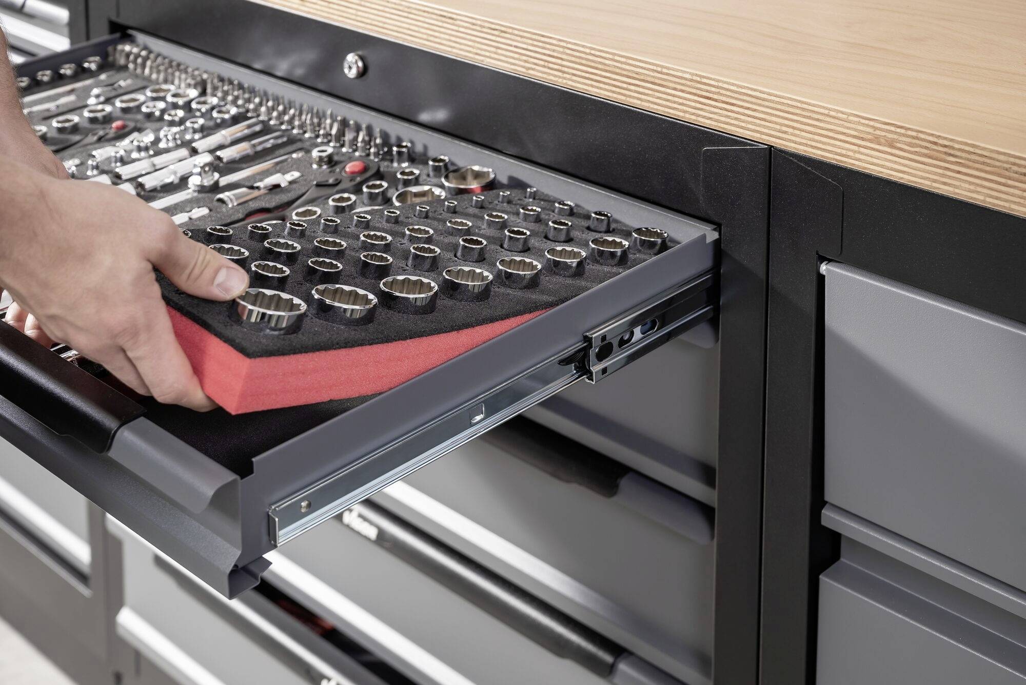 Extendable tool drawer with sorted socket wrenches being pulled out by one hand. Workbench with wooden top in the background.