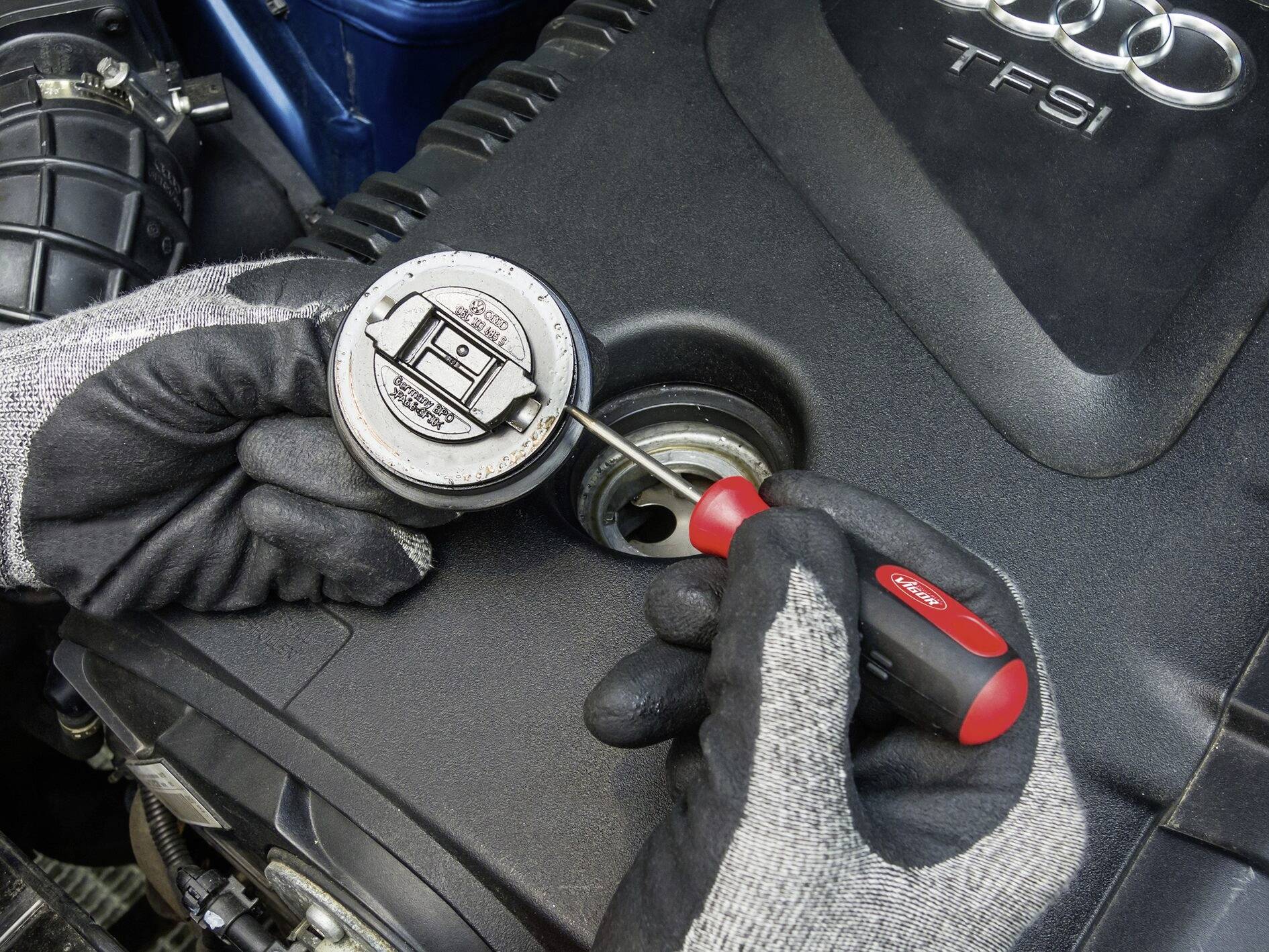 A person wearing gloves opens an oil filler cap marked 'TFSI' using a screwdriver on a car.
