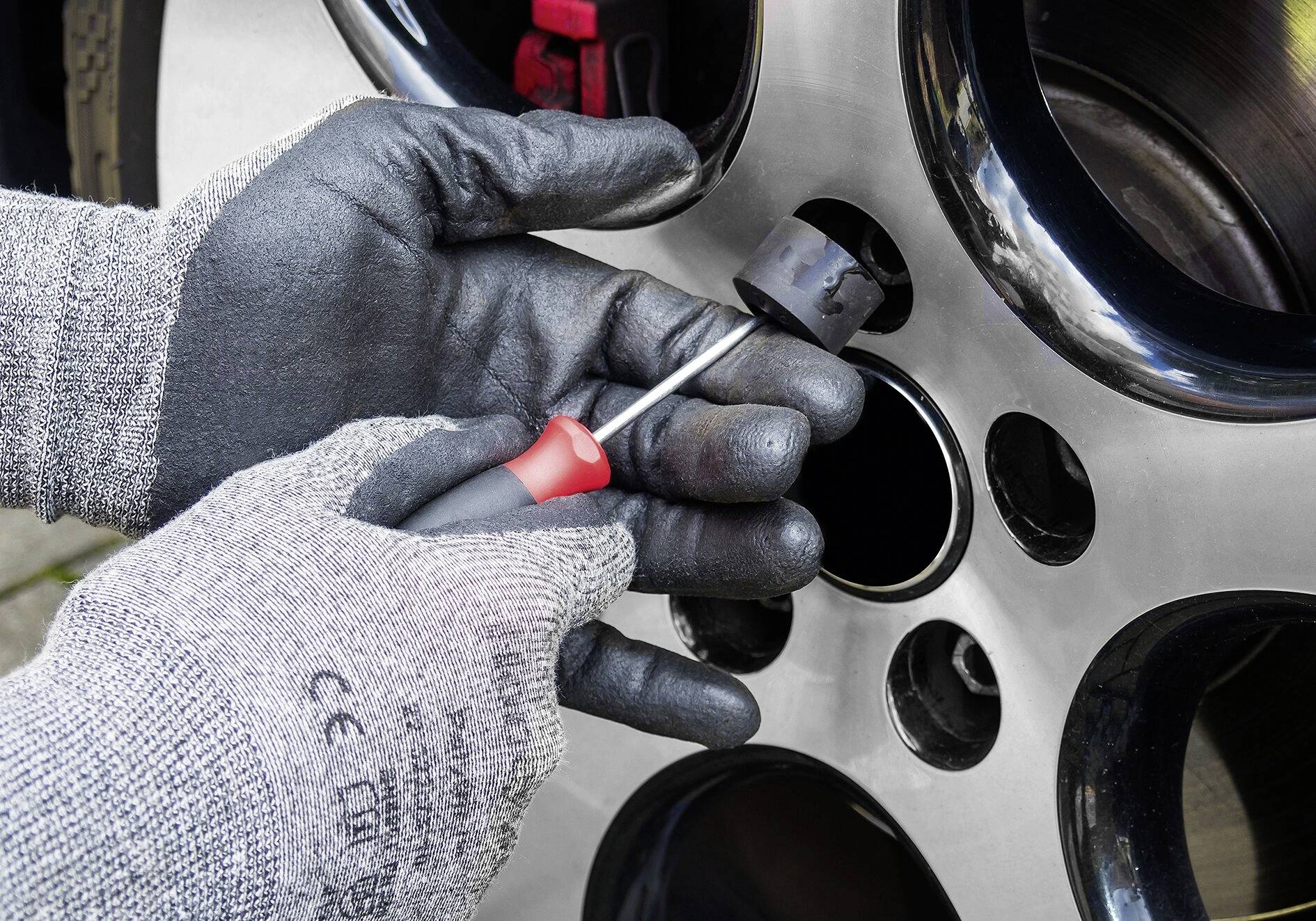 A person is mounting a car tyre with a screwdriver. Hands are wearing black, grippy gloves. A close-up shows the wheel nut and rim.